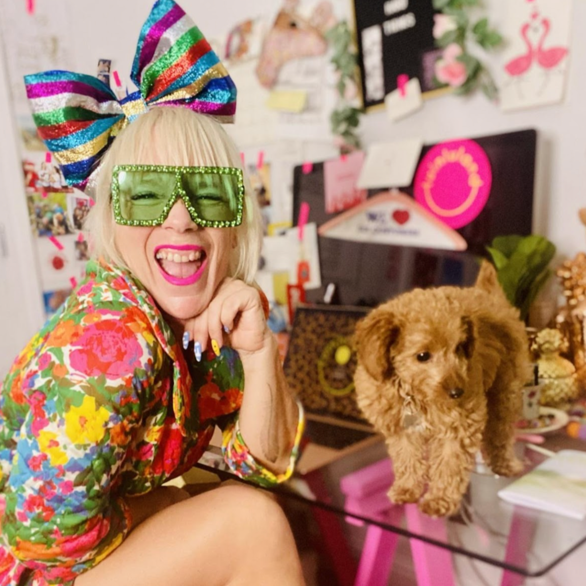Lu is white woman with blonde hair cut into a bob with a fringe. She wears a glitter striped big bow on her head and green sunglasses. Her top is patterned with flowers and she is smiling with her mouth open. There is a puppy on the desk next to her.