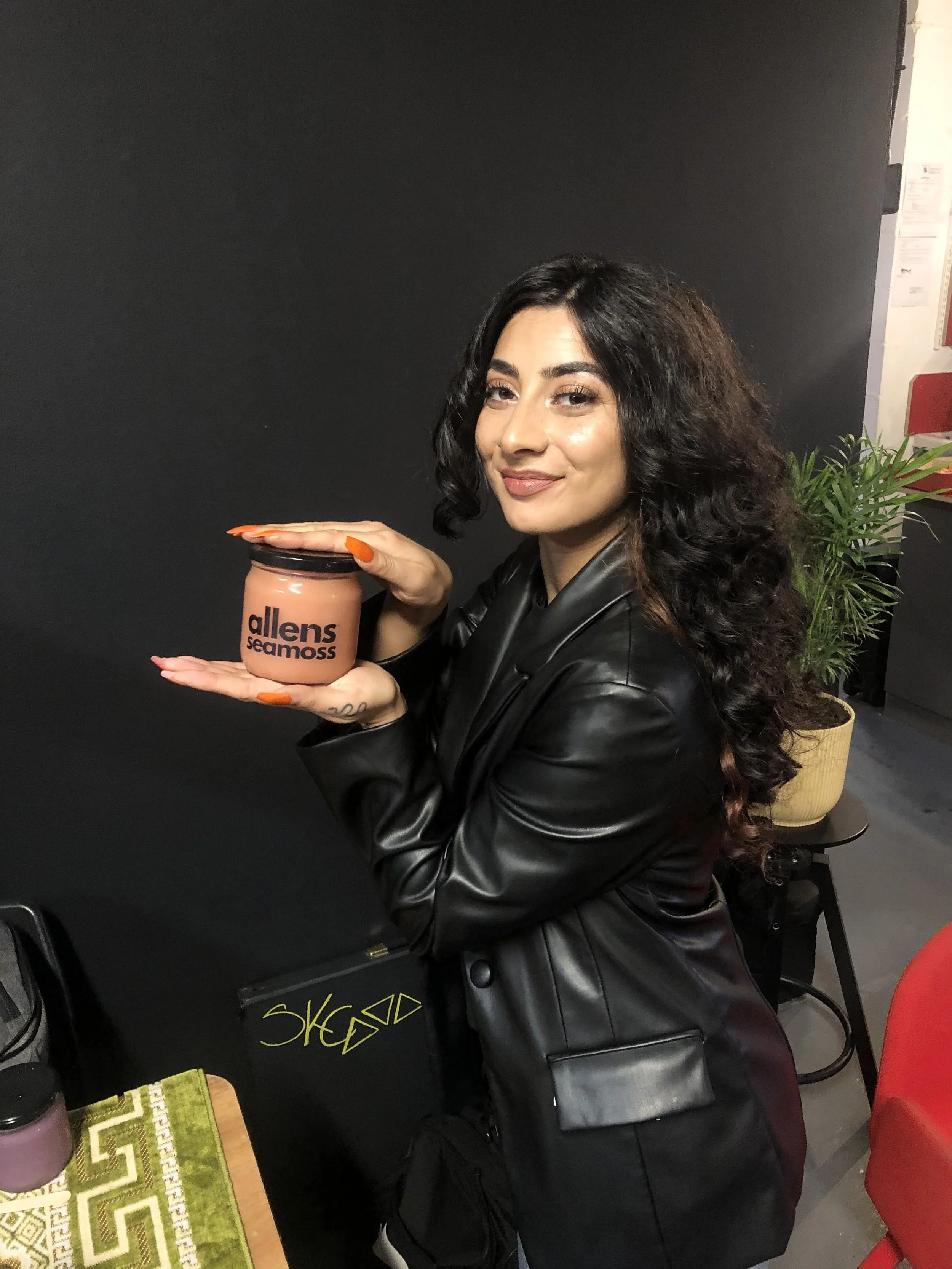 In this close-up shot, a woman (Cristina) with voluminous dark curls holds up a jar of “Allen’s Seamoss” with pride. Dressed in a sleek black leather jacket, she exudes cool confidence.