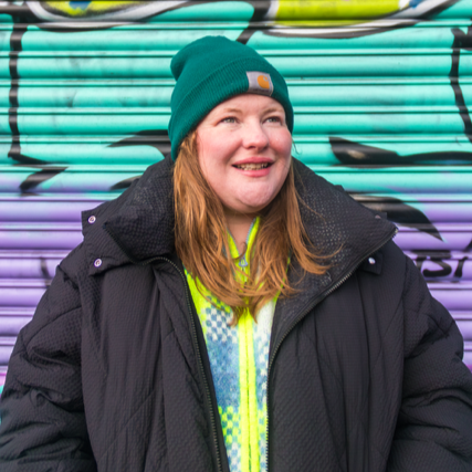A colour photo of Judi standing in front of a multicoloured graffiti shutter. She is wearing a black puffer jacket and a teal beanie, looking off to the side and laughing.