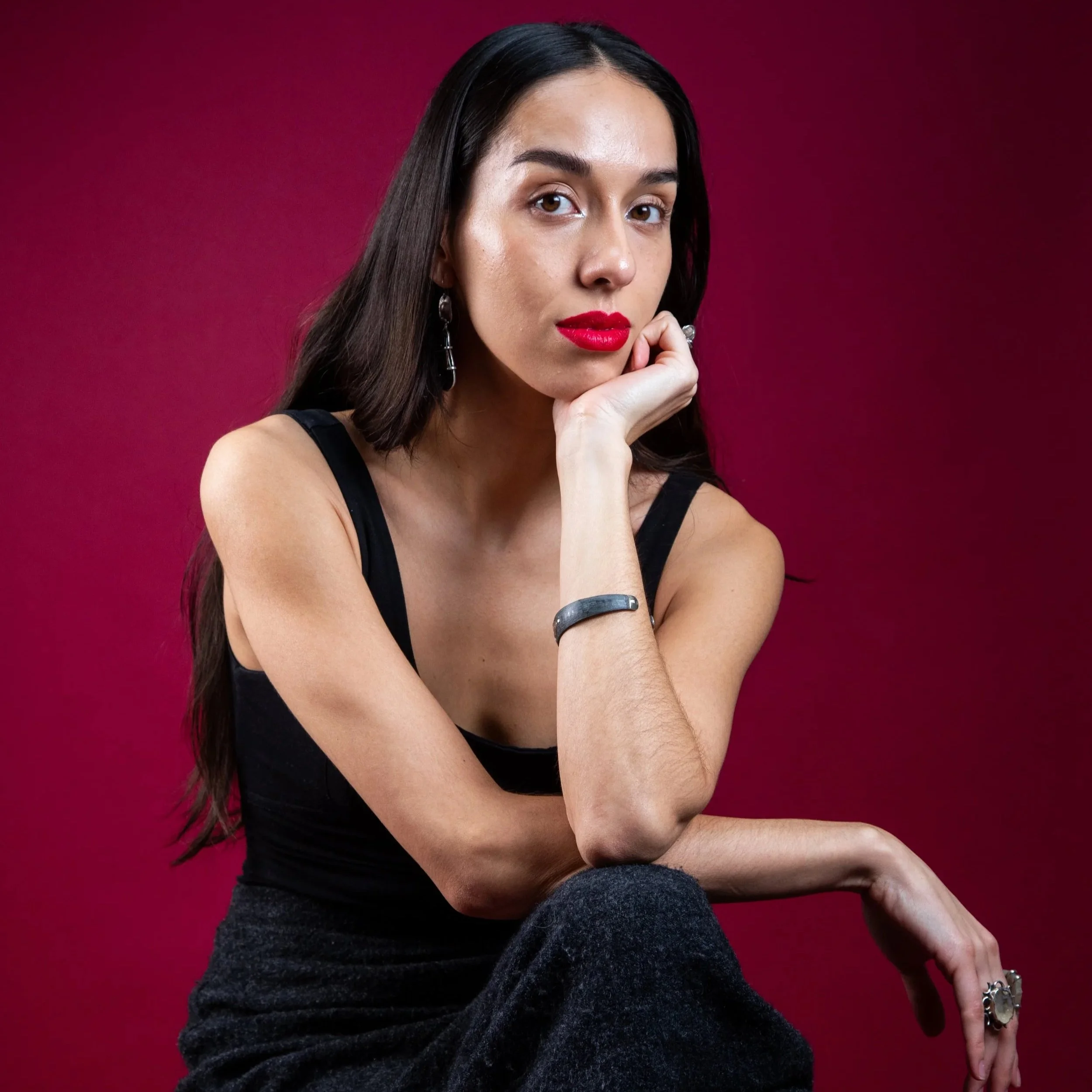 Claudia is a British Mexican woman with long dark straight hair. She wears bright red lipstick, a black vest and skirt. She's sat with one arm over her knee and the other hand placed under her chin