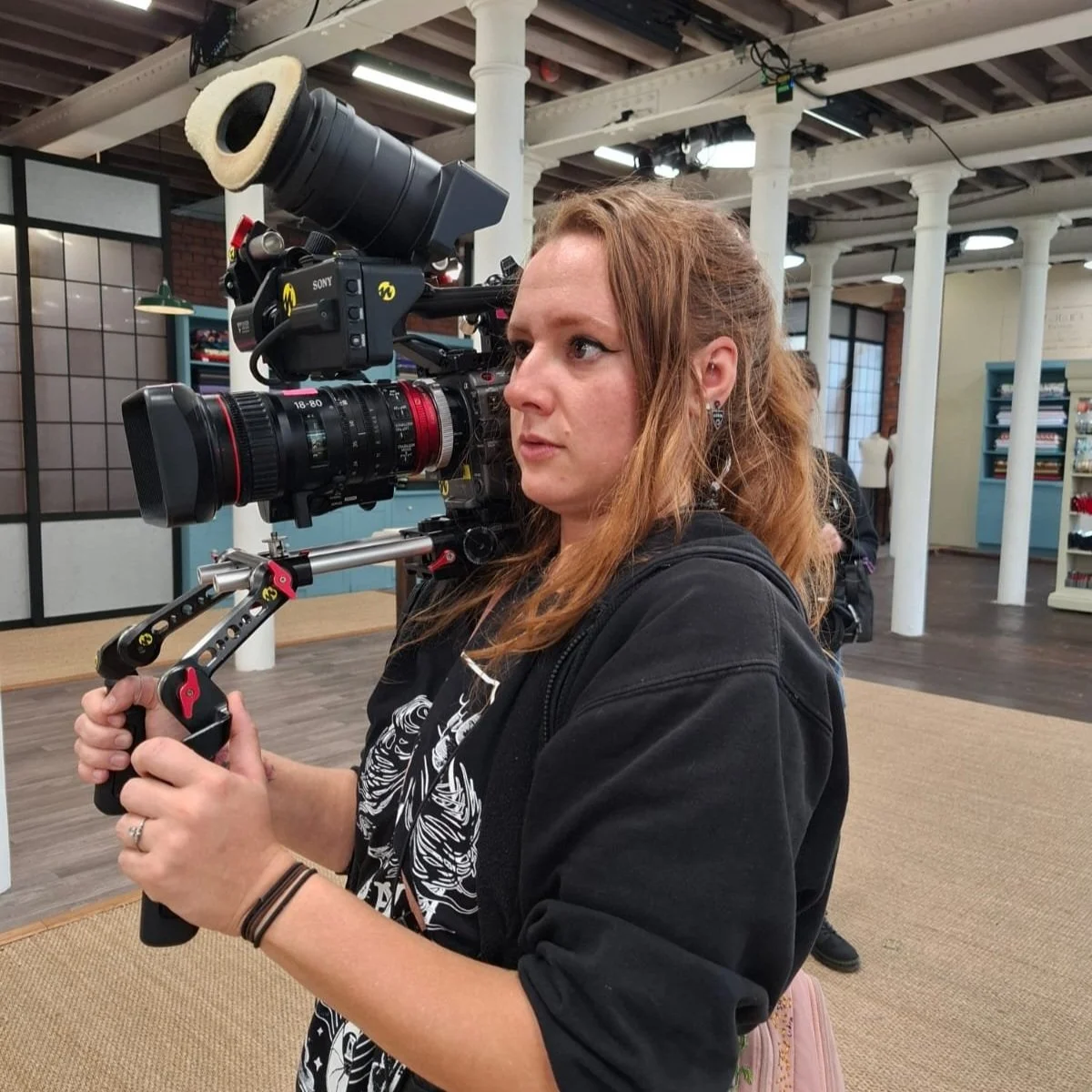 A white woman wearing all black is holding a hand-held camera on set