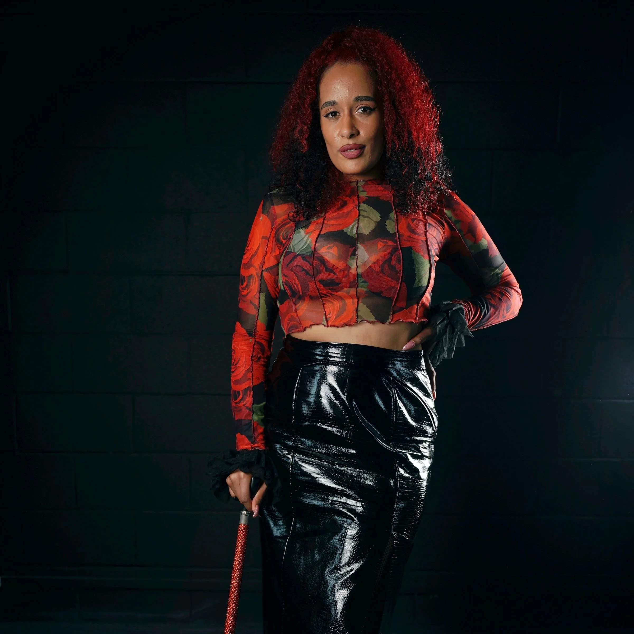 Roxy is a Disabled, Pansexual , Femme standing with their walking stick. They are wearing a black leather skirt and red top