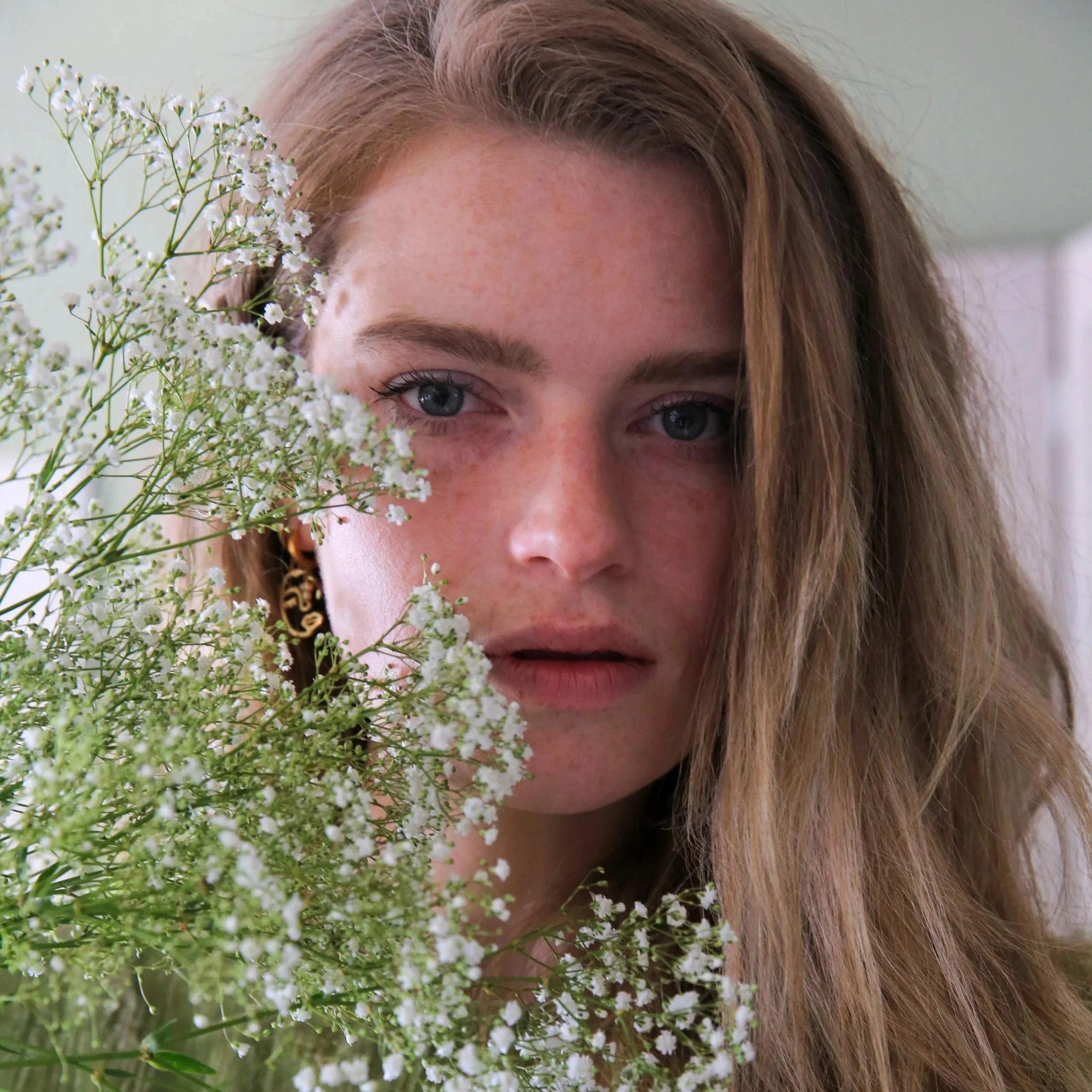 Megan is a white woman with long blonde/brown hair and blue eyes. She is peering behind green and white foliage.