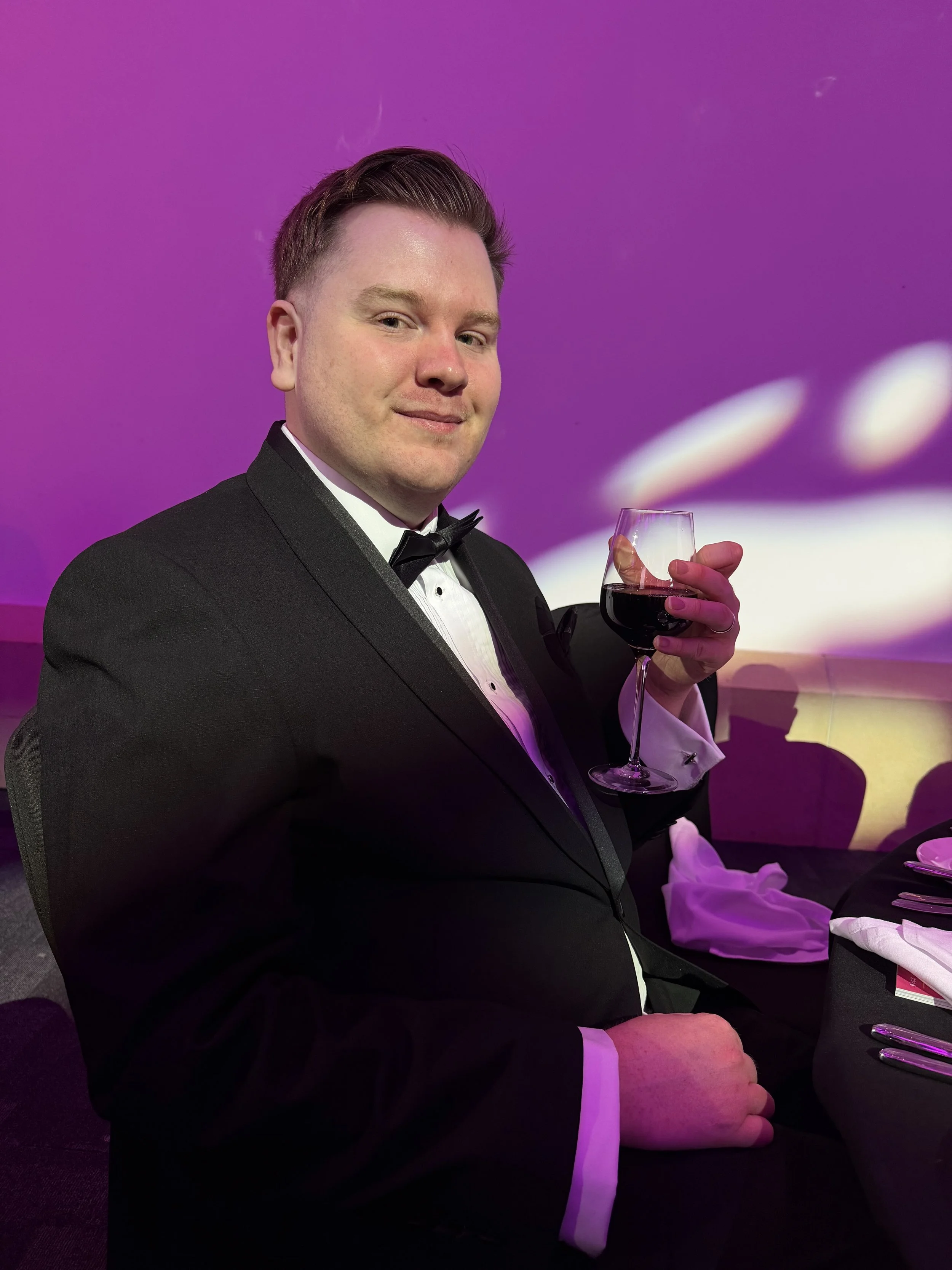 Nicholas sat down with a glass of red wine at the Welsh BAFTAs. He's wearing a black suit, white shirt and bowtie in front of a purple background