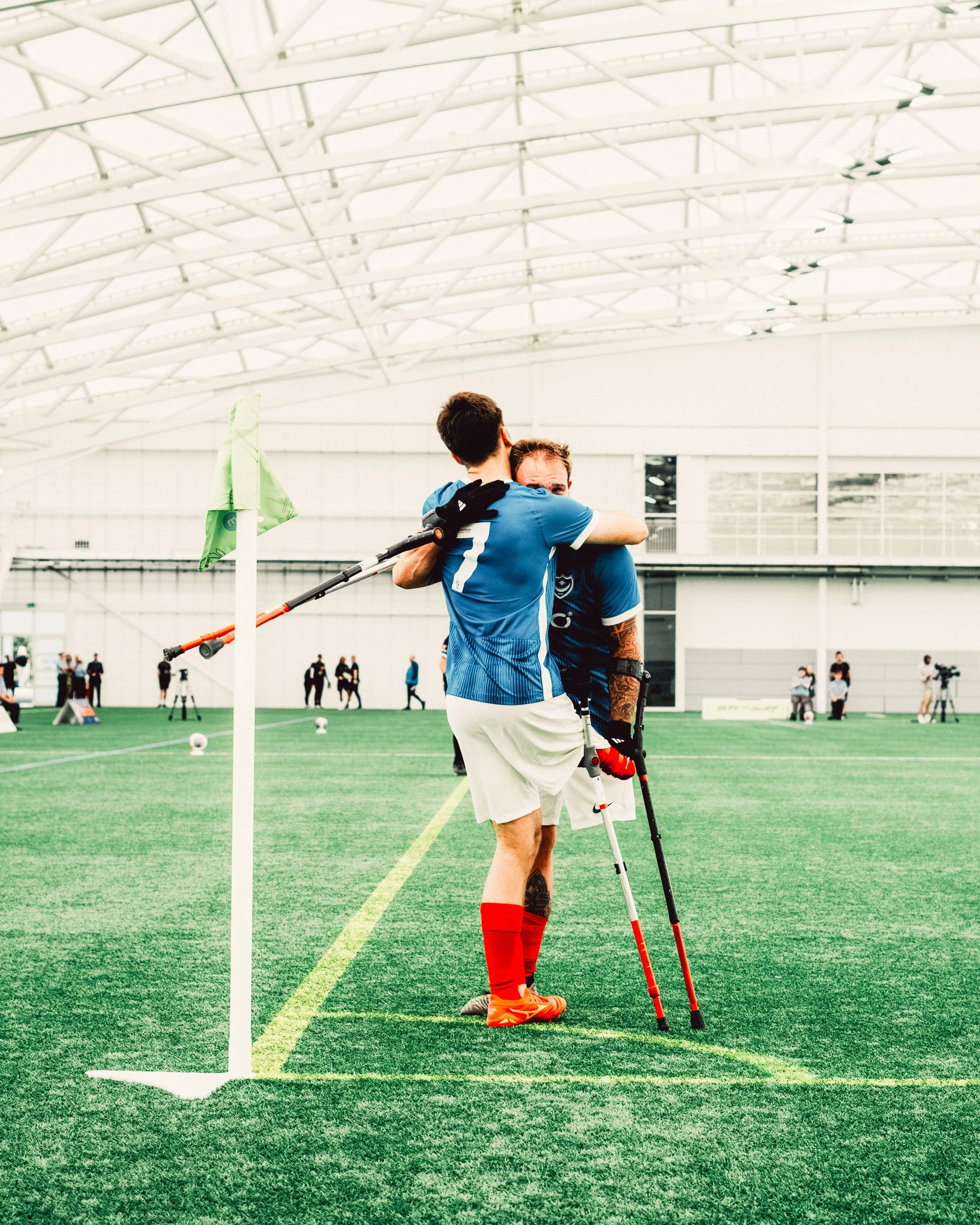 Two athletes hugging on an indoor pitch. They are both white men and each have lower limb difference and use crutches.