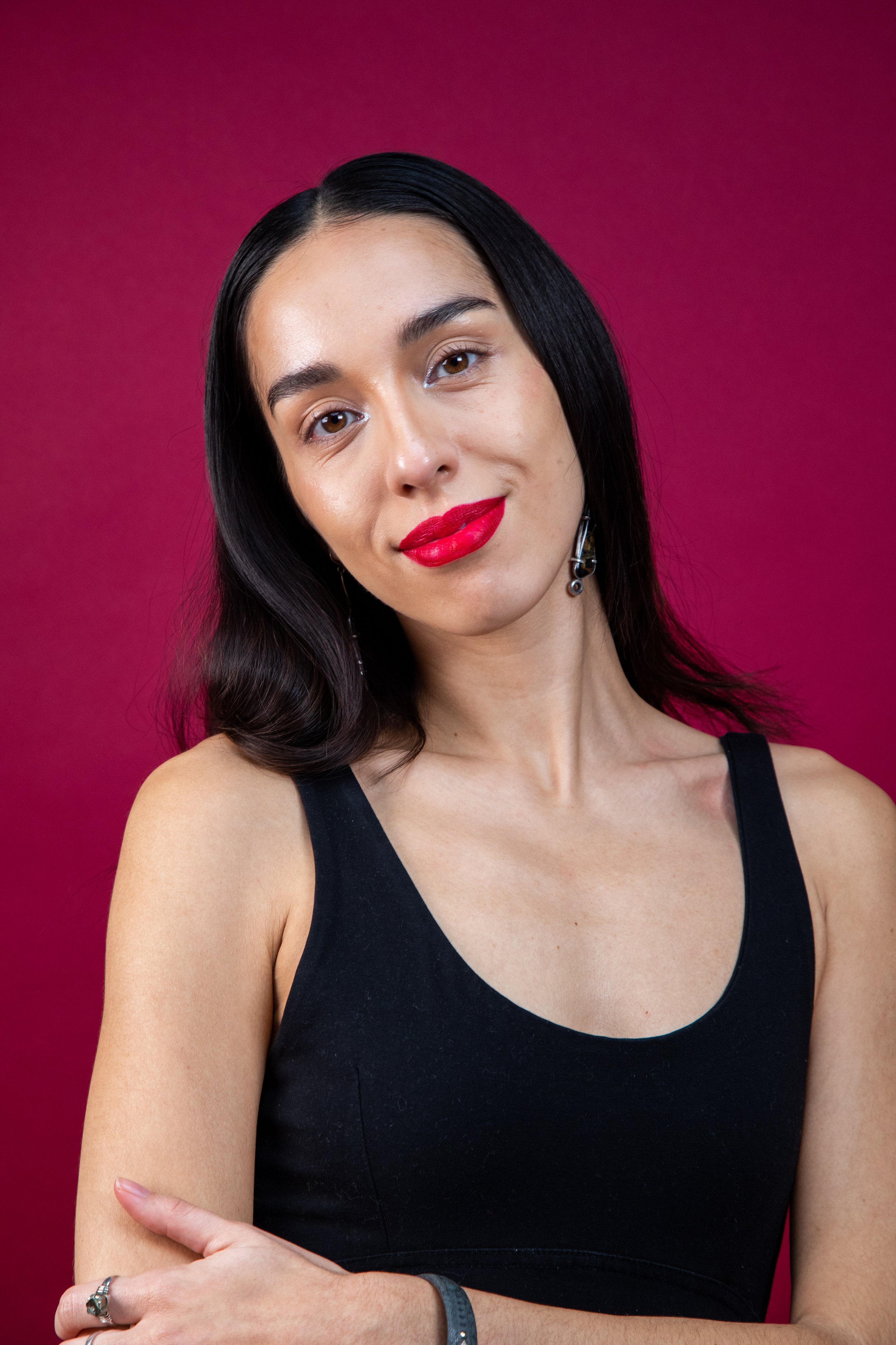 Claudia is stood against a red backdrop with her arms crossed over her body in front of her. Her hair is long, dark and parted in the centre. She wears red lipstick and a black vest