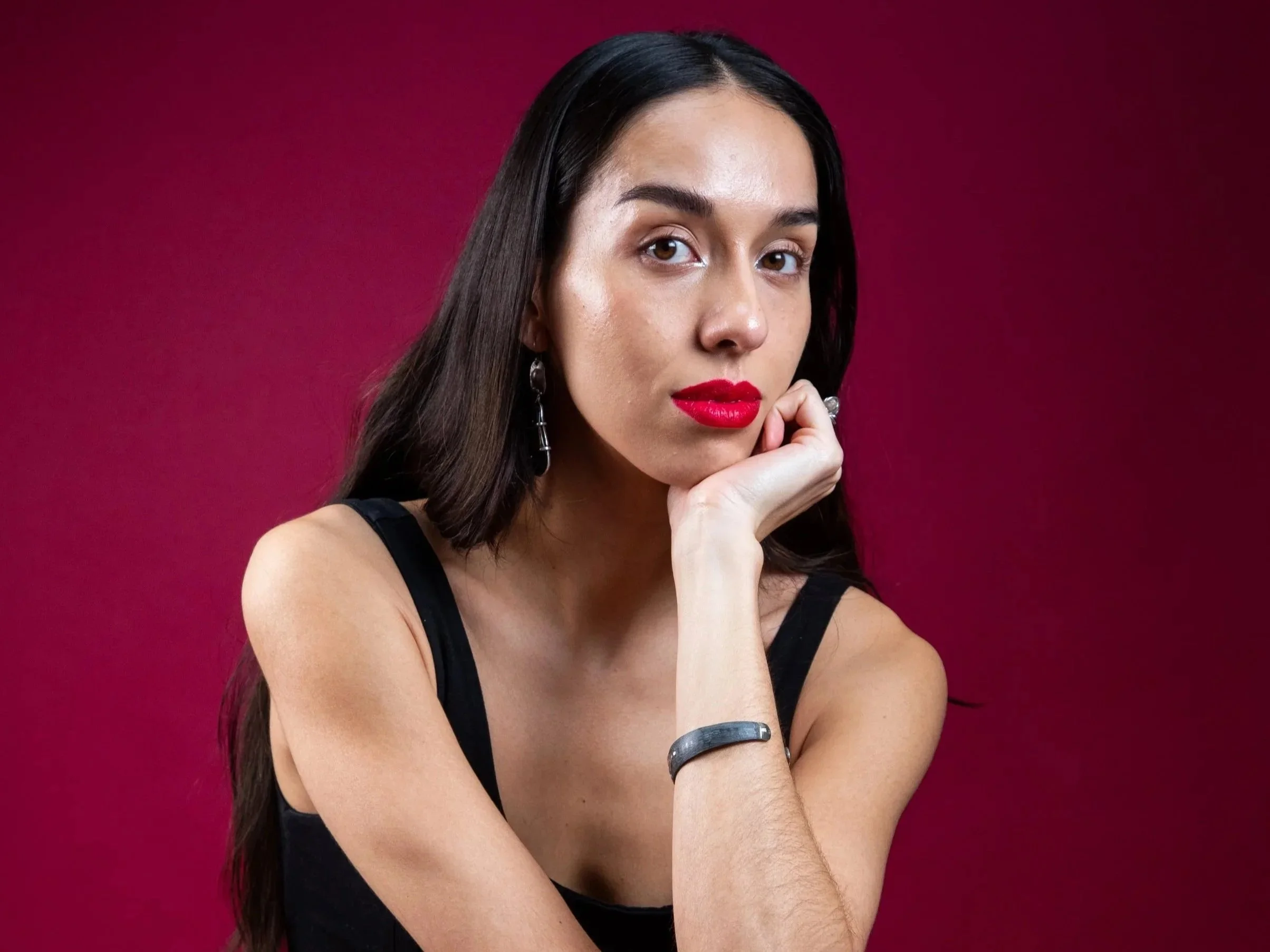 Claudia is latinx with long dark hair parted in the middle. She looks at the camera with her chin resting on her hand, wearing bright red lipstick against as red background.