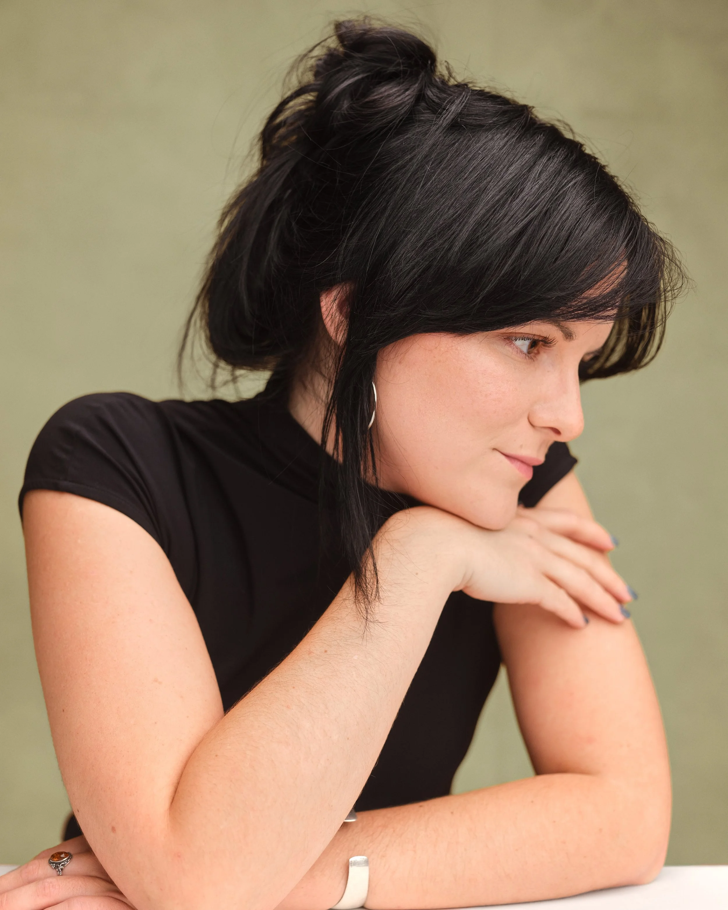 Abbie is wearing a black sleeveless top with her hair styled up in a clip. She has her arms folded and is resting her chin on her hand as she looks to one side.