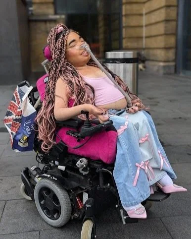 Shelby in London wearing denim flared jeans with bows on them, with a matching pink crop top and pink sparkly heels. Her pink braids are half up half down. She is wearing natural makeup.