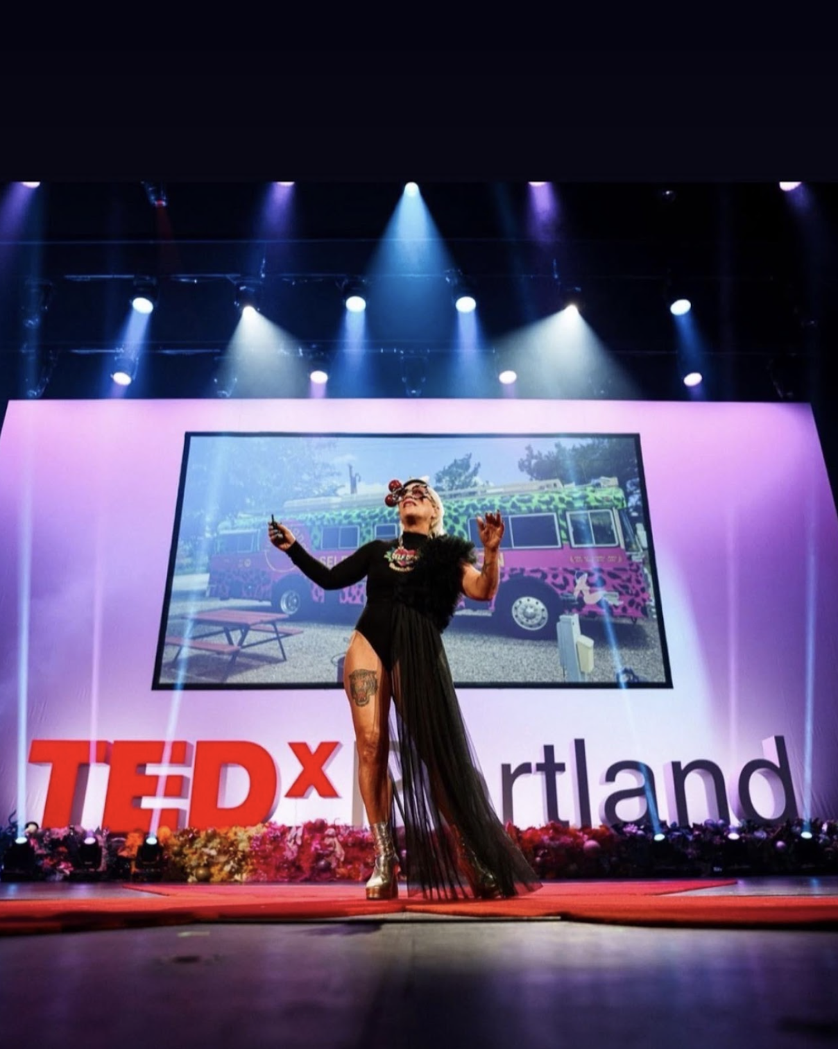 Lu is on stage at her TEDx talk in Portland. She's stood in front of the screen with her arms out wearing a long black dress with a thigh high slit up on side. She had tattoos on her leg and wears silver platform boots