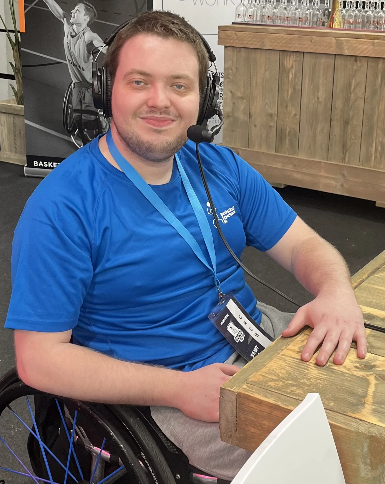 Dylan sits in his wheelchair at a table wearing a blue t.shirt and lanyard with a commentator headset.