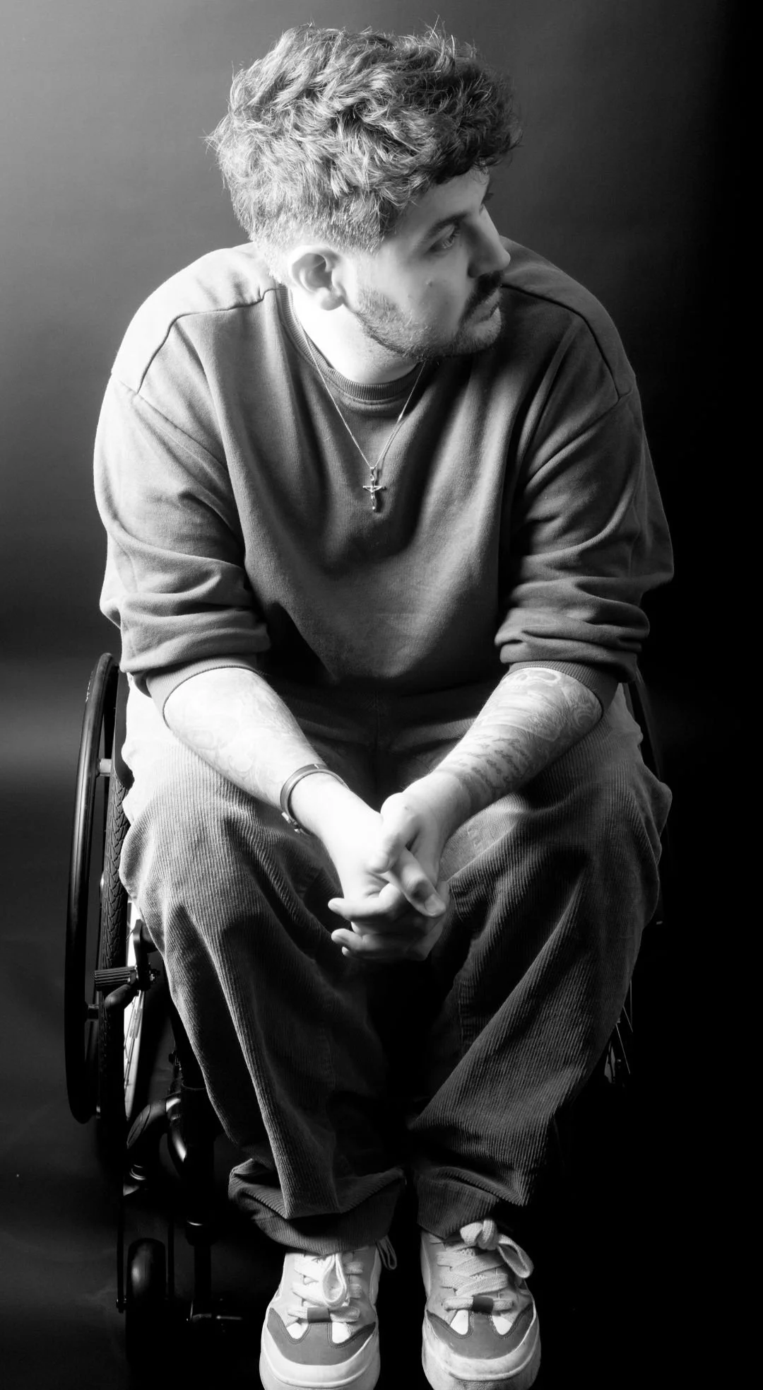A black and white image of Nathan sitting in his wheelchair looking to the side. His hands are clasped in front of him, resting on his knees.