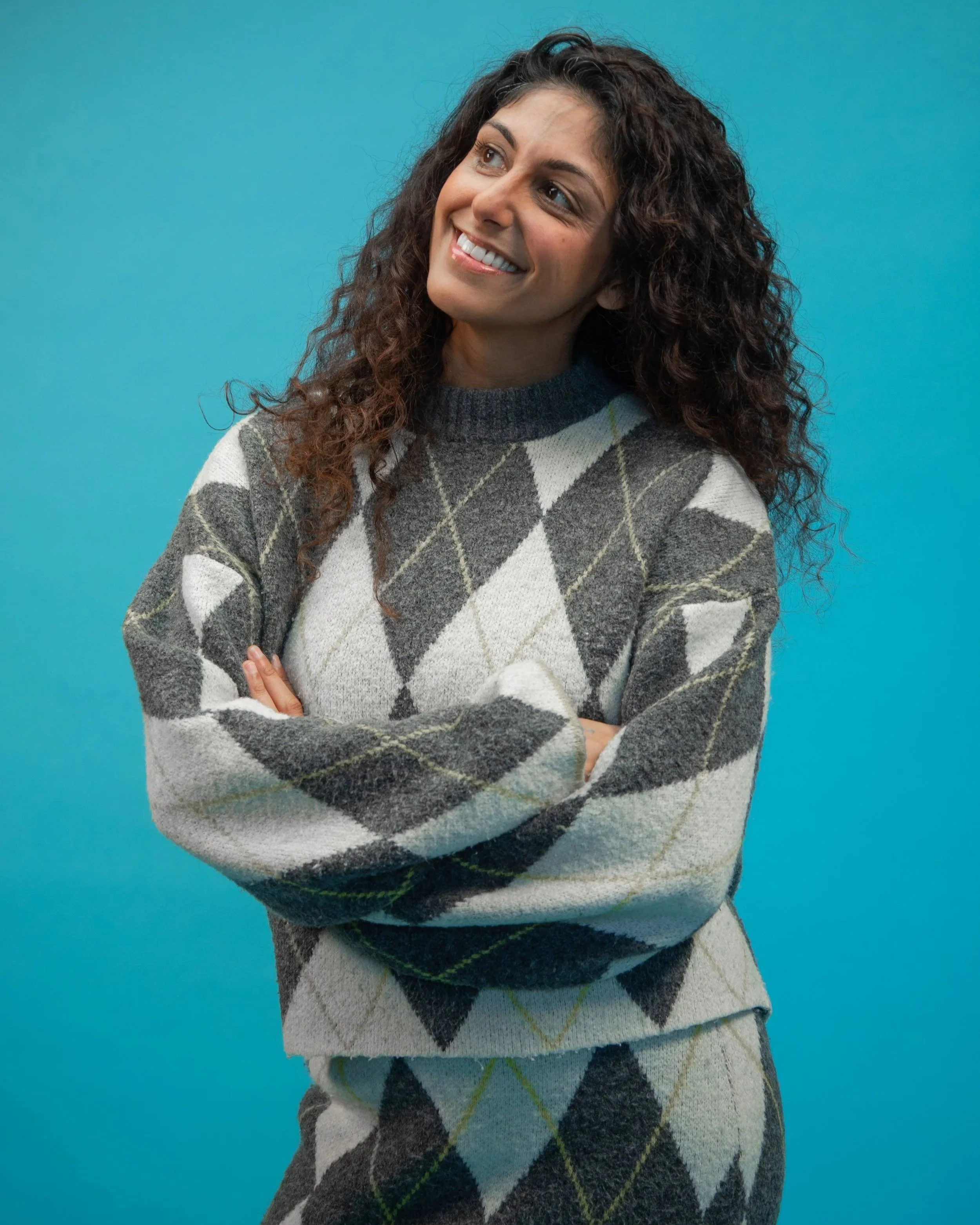 Amanprit is looking to the side smiling with her arms folded in front of her. She wears a matching grey knitted skirt and jumper.