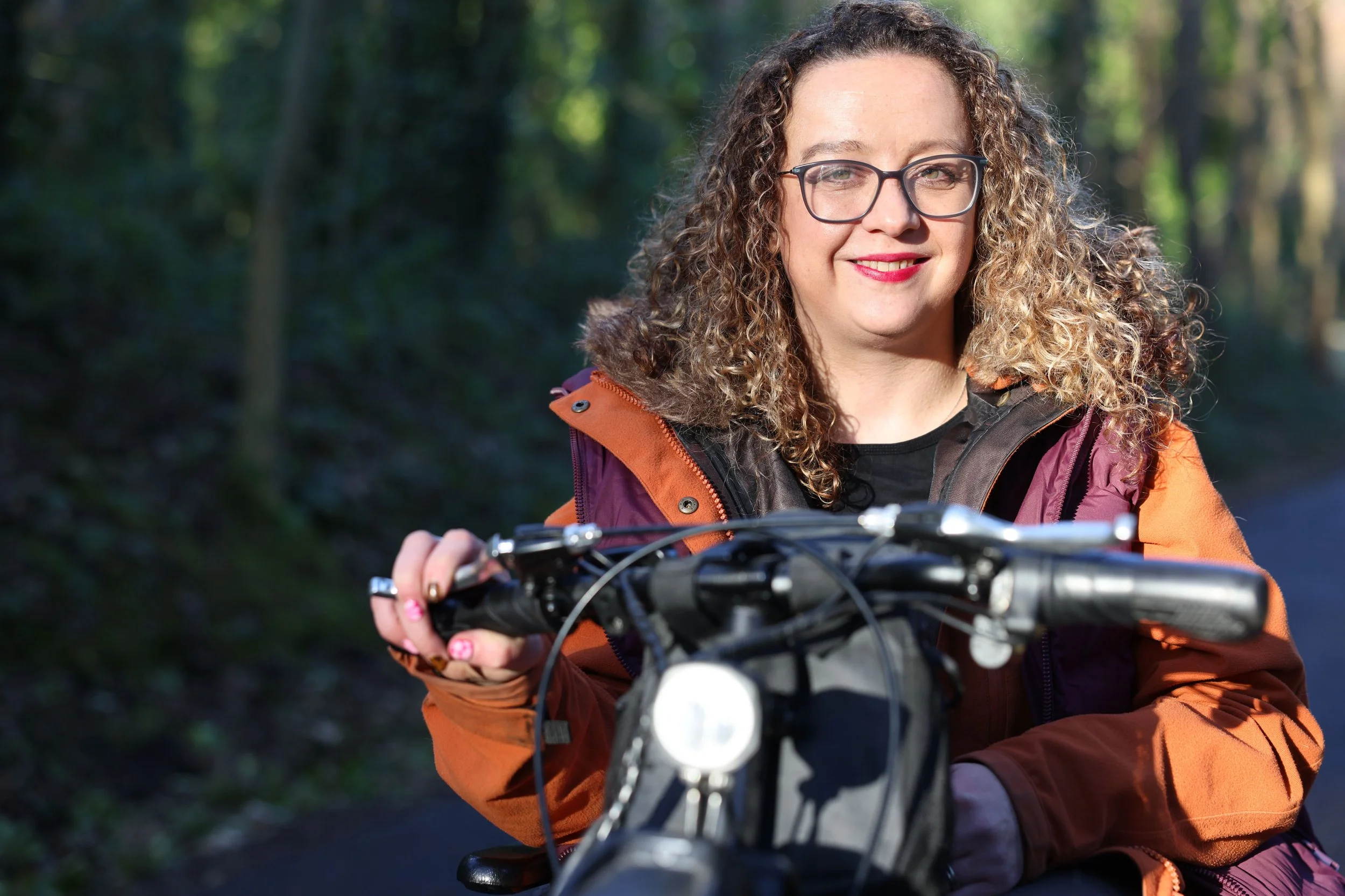 A woman with curly hair and glasses is outdoors in a wooded area, holding the handlebars of a handcycle attachment on her wheelchair. She is smiling and wearing the same two-tone jacket.