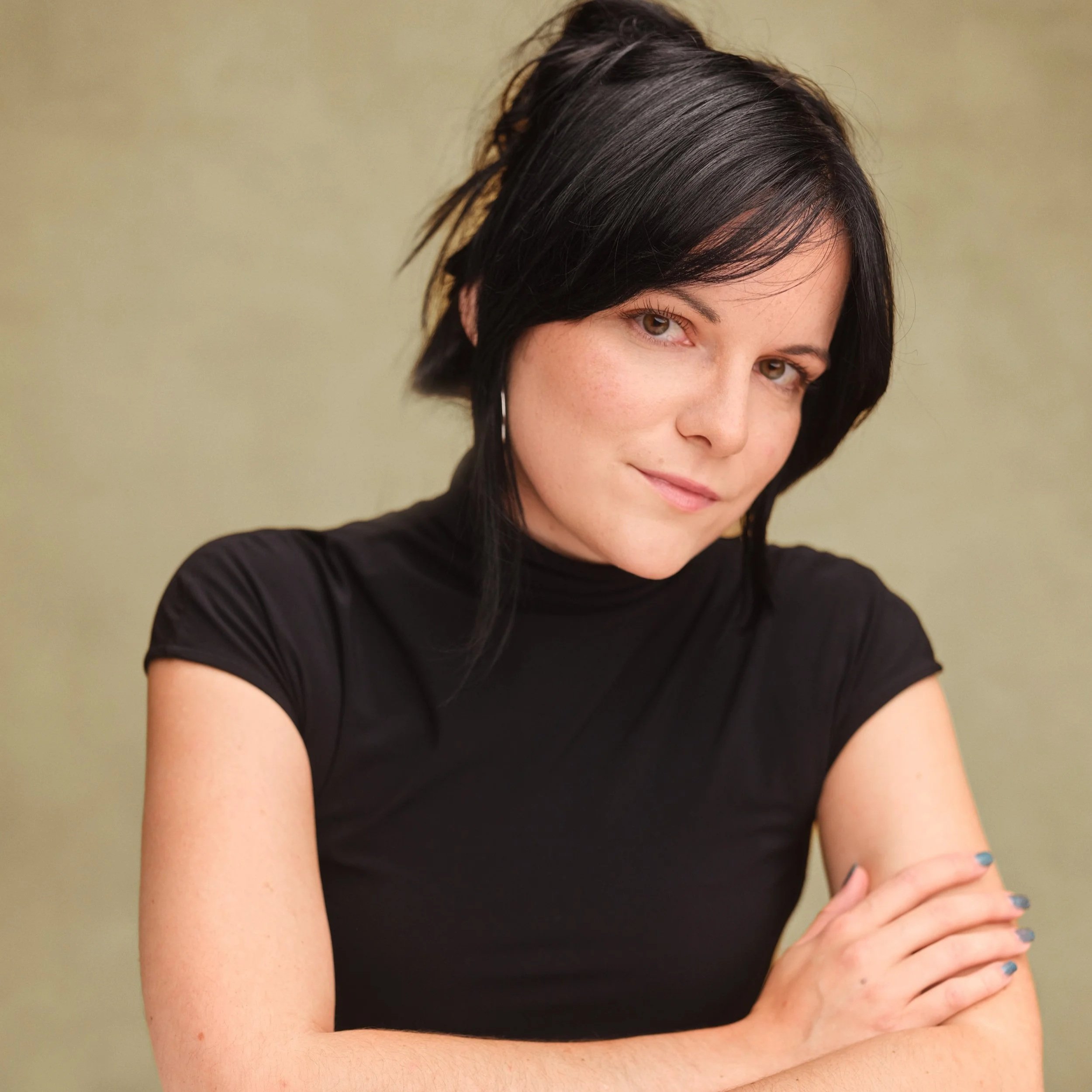 Abbie is a white woman with dark hair styled up in a clip. She is sitting at a table with her arms folded, wearing a black top