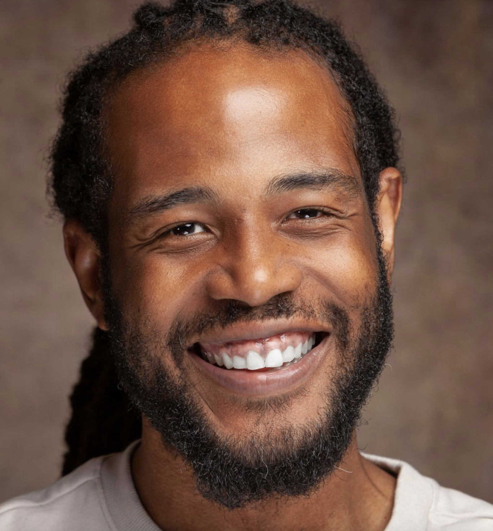 Ekow is smiling a the camera with his braids pulled back behind his head. He's wearing a white t.shirt against a brown background