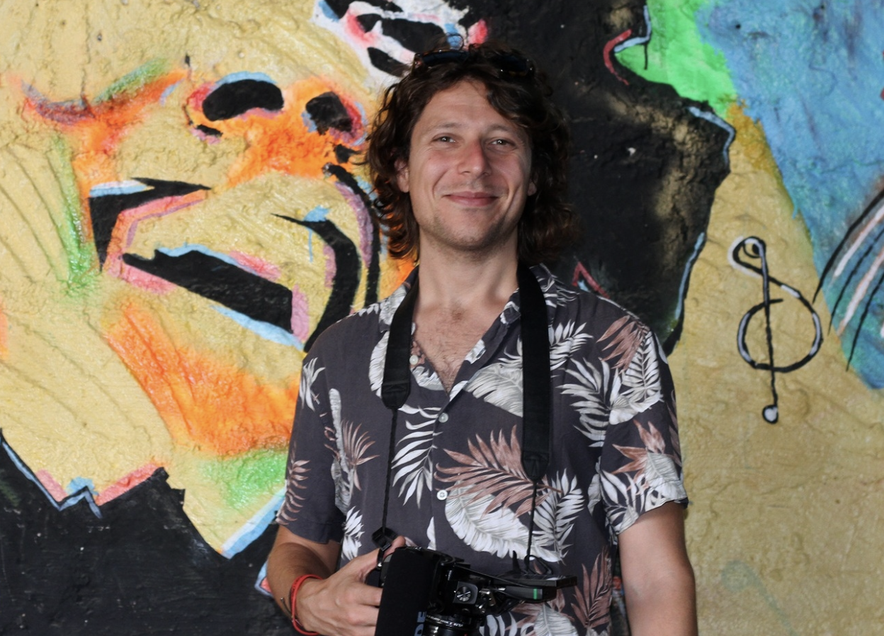 James is a white man with brown curly hair. He's wearing a patterned shirt and is holding a camera around his neck