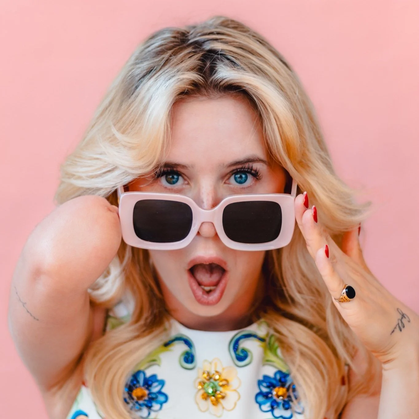 Brooke is a white woman with blonde hair. She has upper limb difference in her right arm and is peering over pink sunglasses.