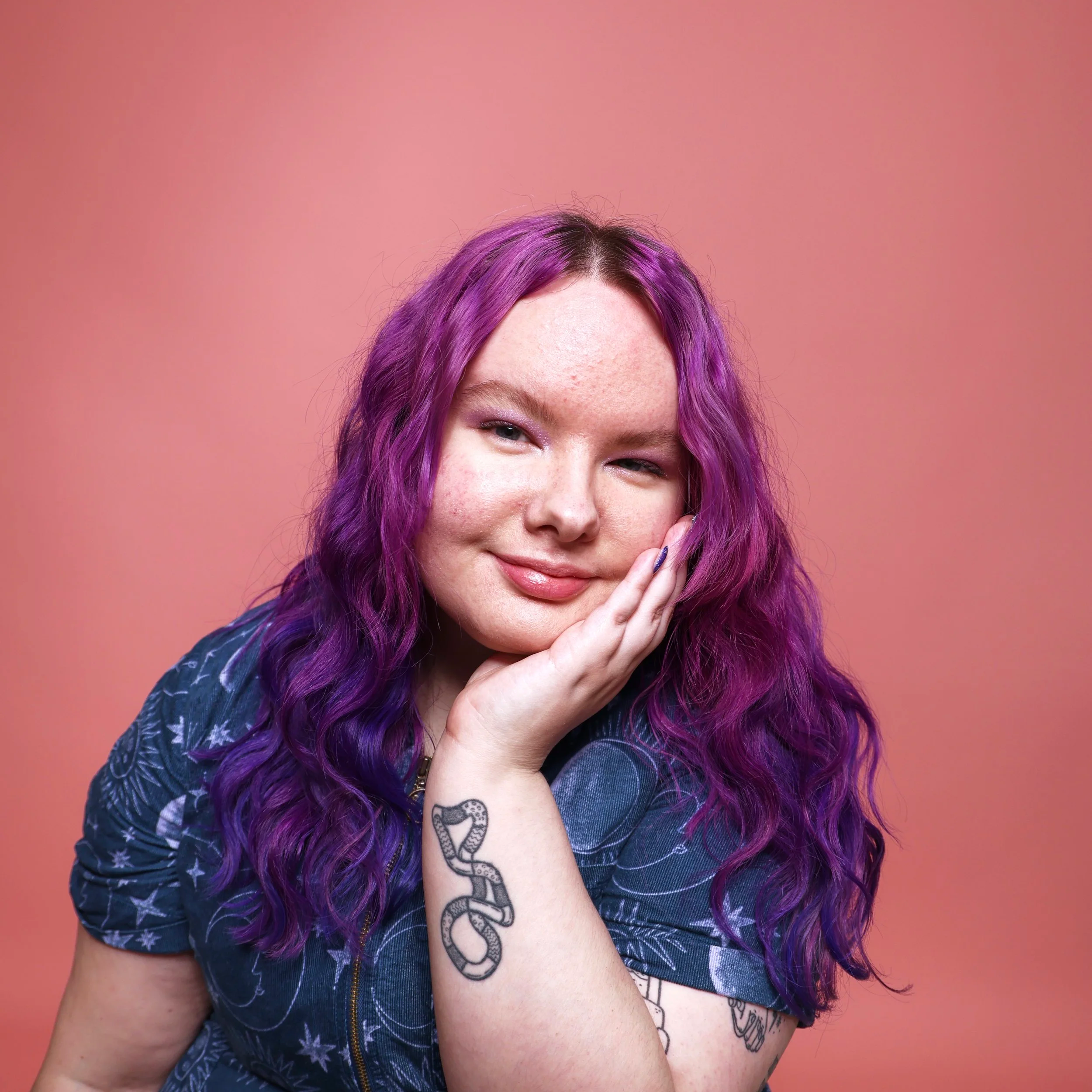 Charli is a white person with purple wavy long hair. They are wearing a blue patterned shirt and have tattoos on their arm. They are resting their face on one of their hands.
