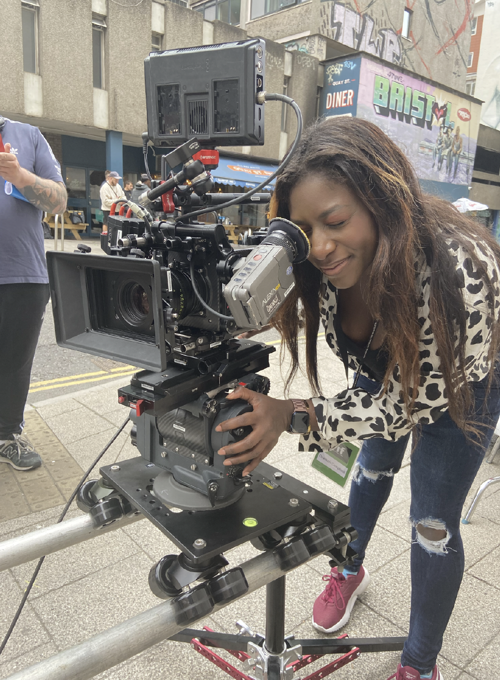 Oneikeh is on set looking into a camera. She wears a leopard print top and jeans with red dark pink trainers. Her hair is long, brown and wavy.