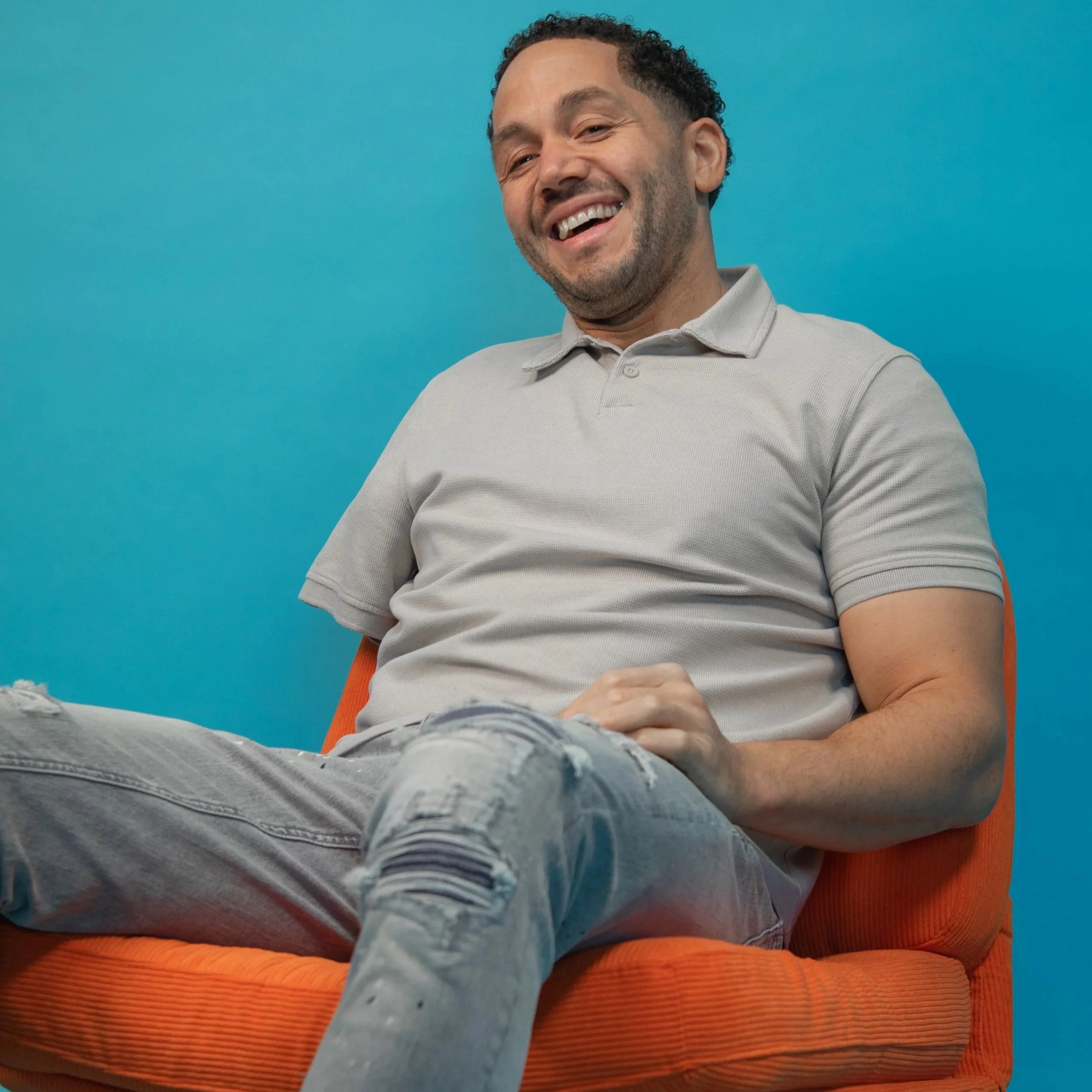 Chris is sat back on the orange chair with his hand on his lap, laughing into the camera.