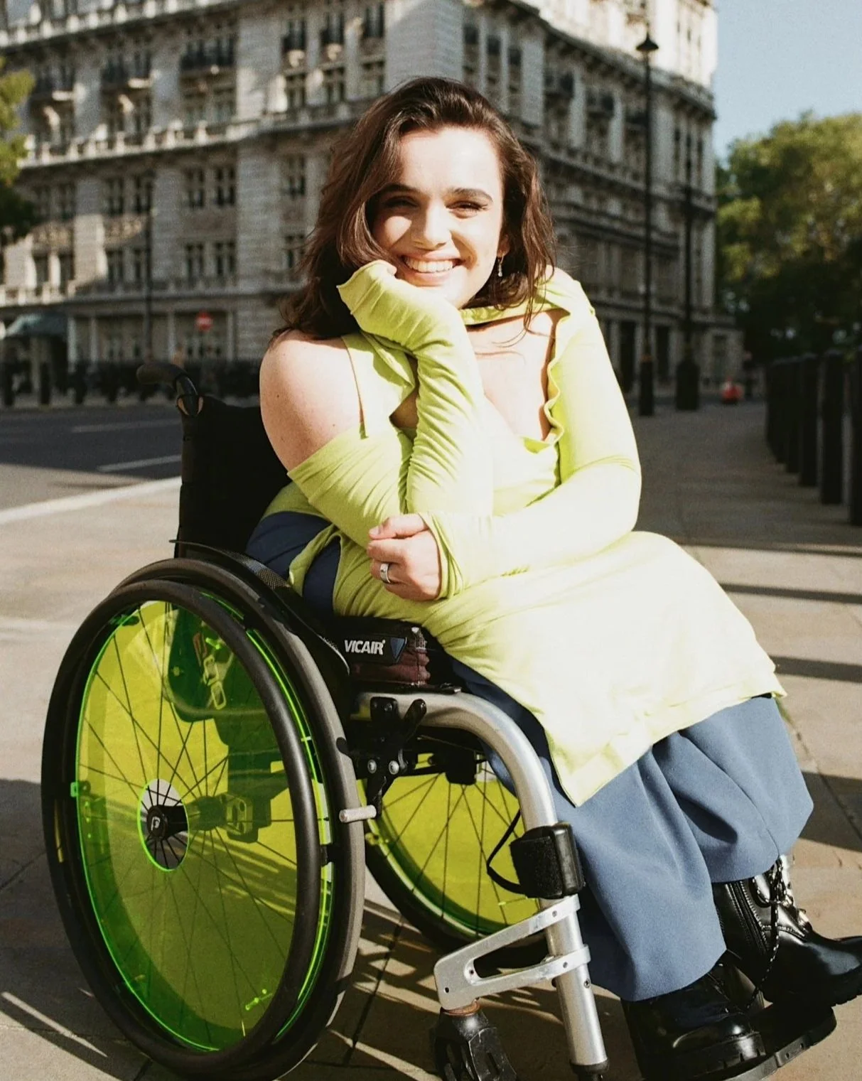 Alexandra sitting in her wheelchair wearing a lime green long top over blue trousers. Her hair is long, soft and wavy and she smiles at the camera with her head resting on her hand.