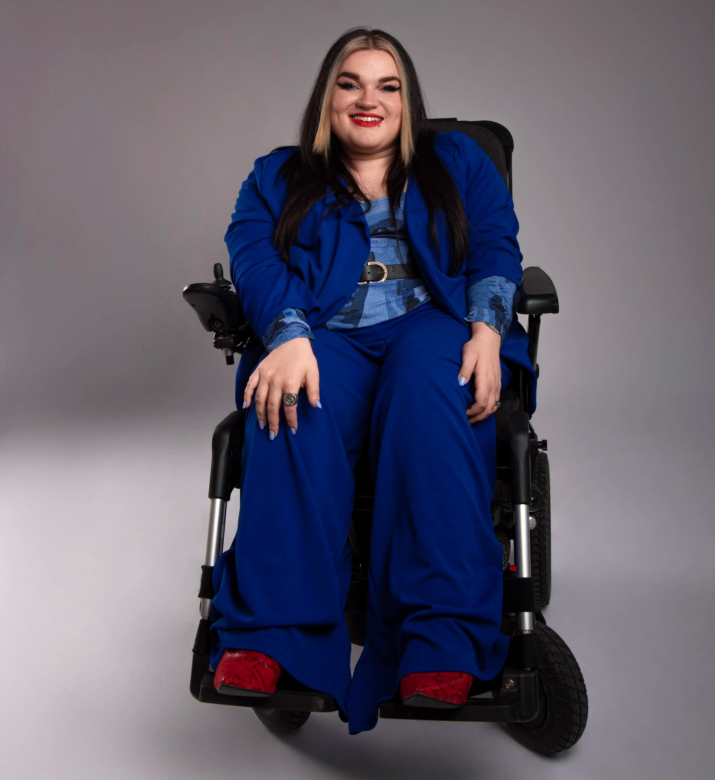 Kelly is a white woman with long dark hair with blonde strands at the front. She sits in her power wheelchair wearing an electric blue suit and red snakeskin boots.