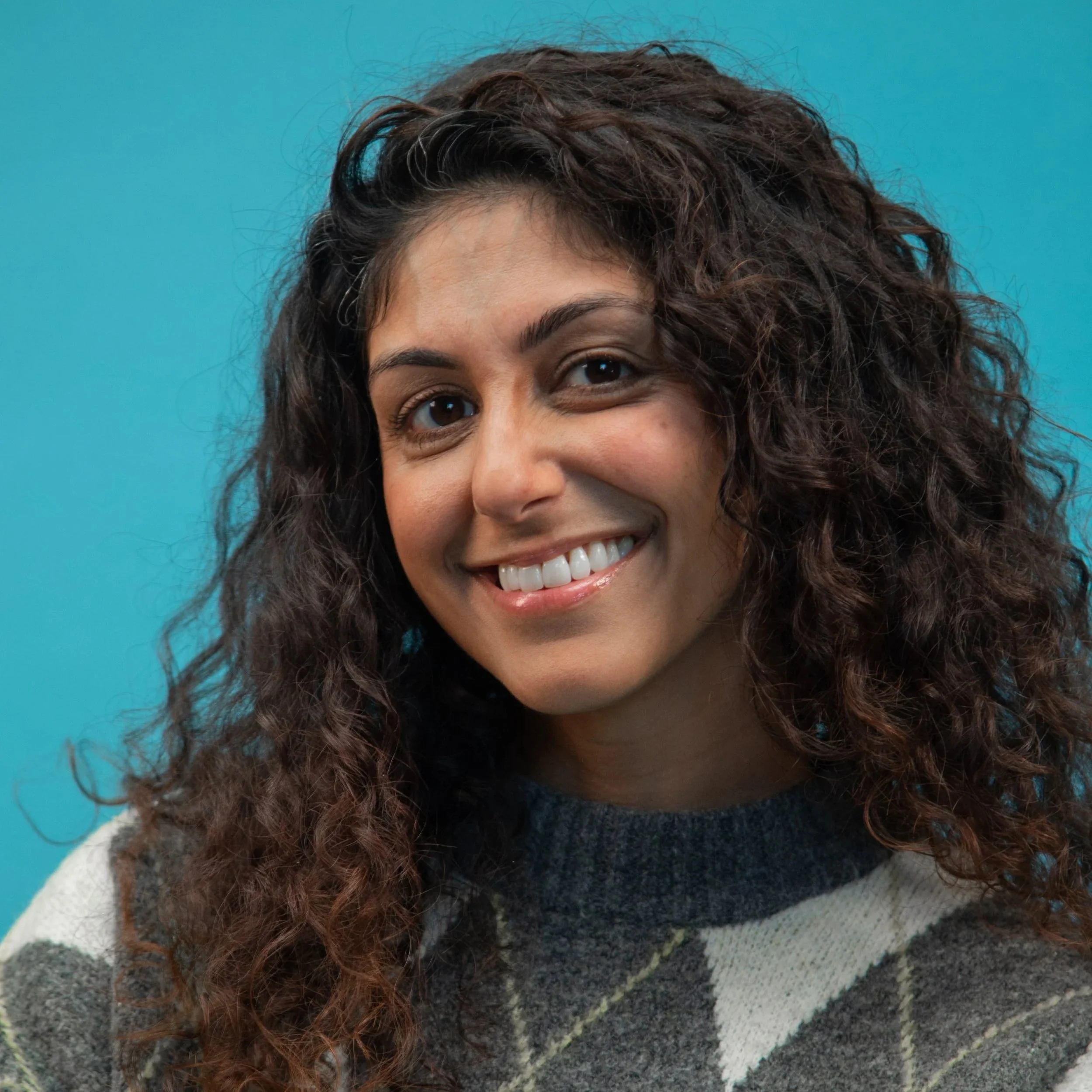 Amanprit is a brown deaf woman with long brown curly hair. She is smiling, looking at the camera in front of a blue backdrop.