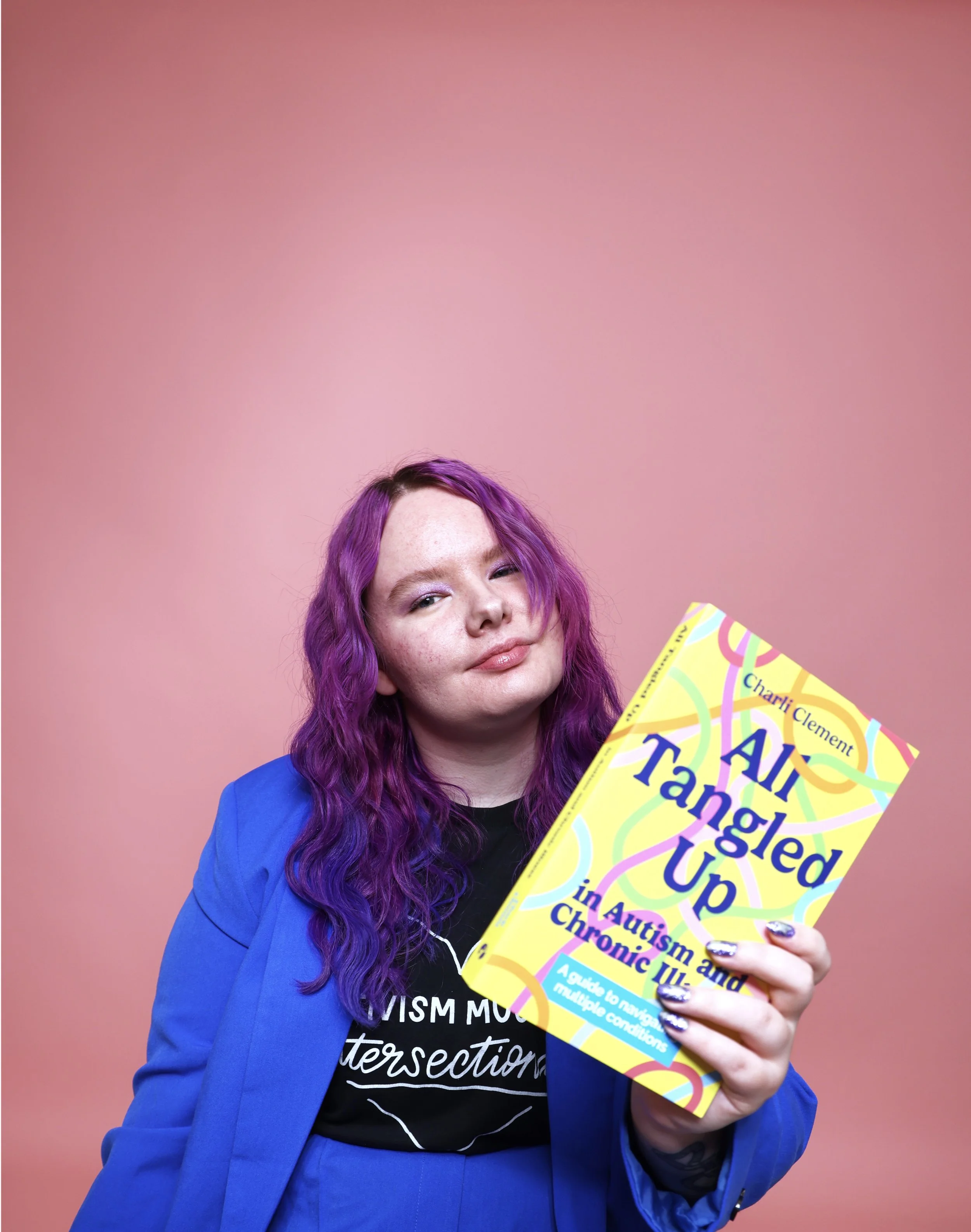 Charli is wearing an electric blue blazer and black t.shirt against a soft pink background. They have long wavy purple hair and they are holding a copy of their book ‘All Tangled Up in Autism and Chronic Illness’.