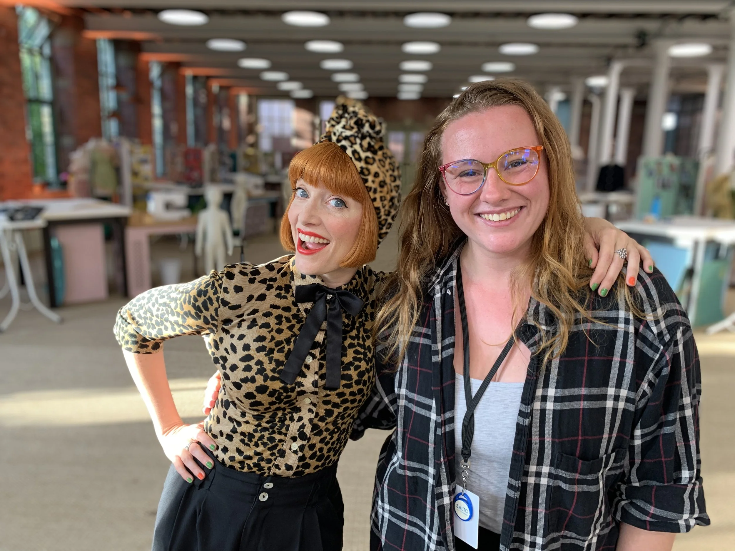 Xenia, a white woman with long hair and glasses, poses on set with Amber Butchart who has bright ginger hair, wearing all leopard print.