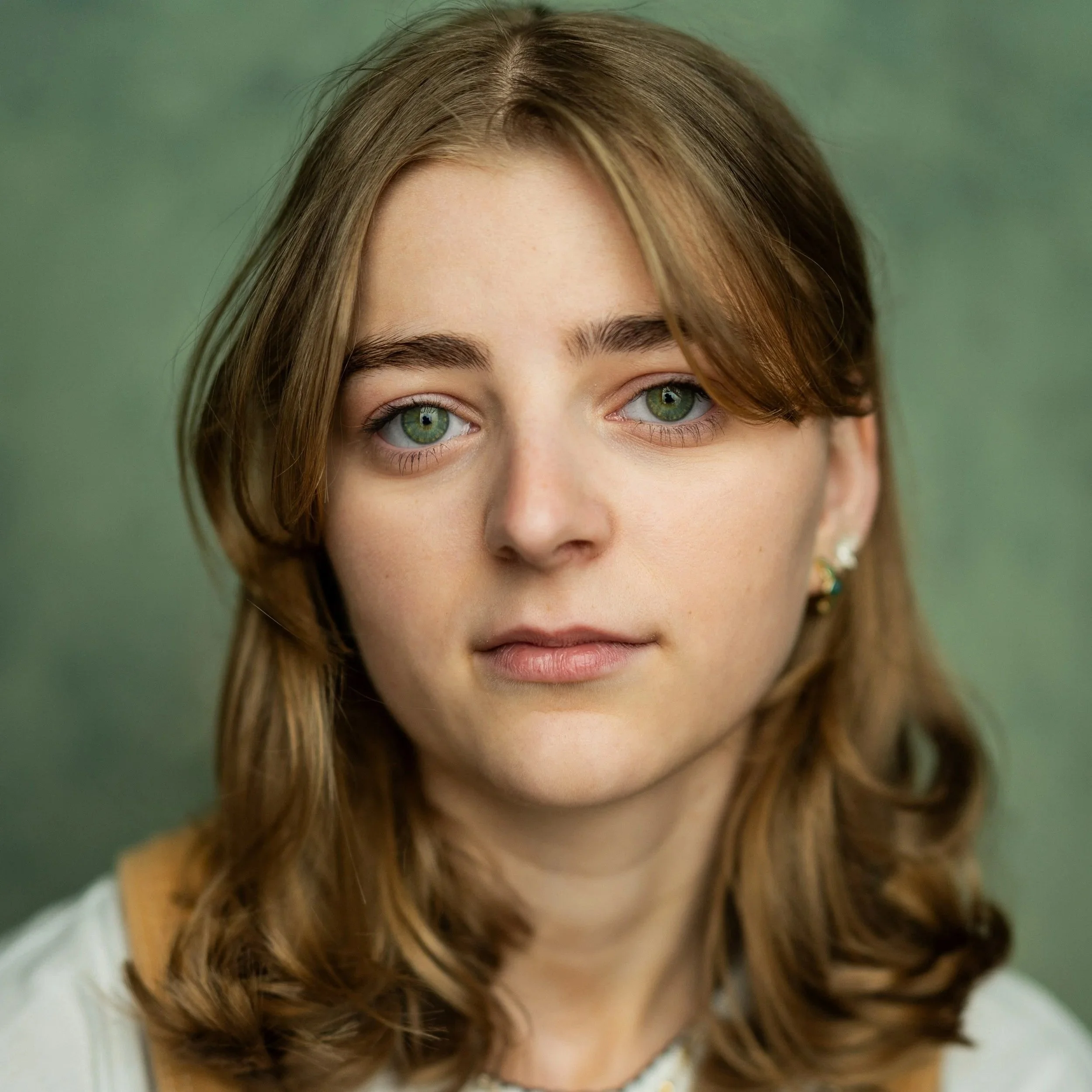 A white woman with long blonde/brown hair, parted in the centre. Her eyes are green and she wears a white tshirt and dungarees