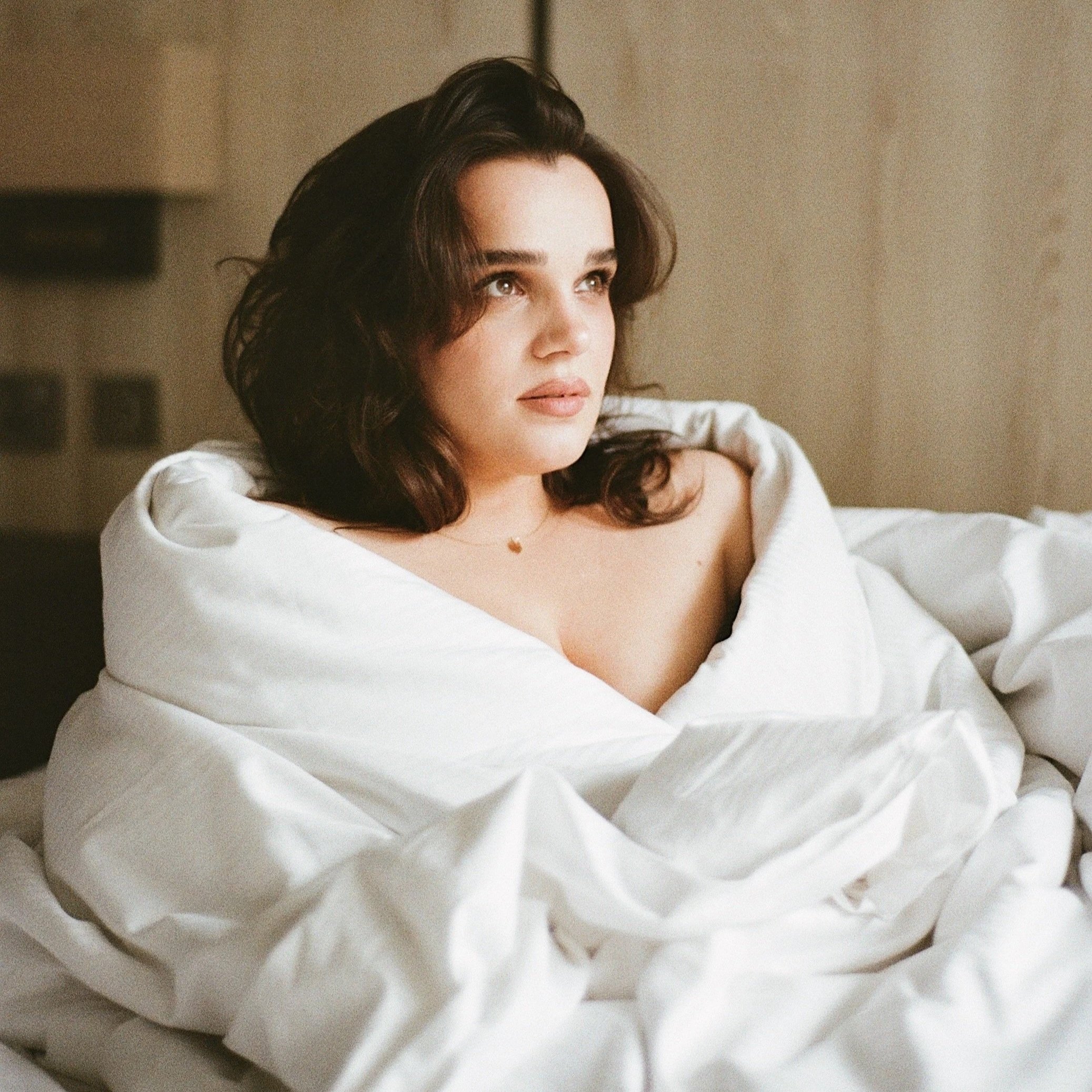 Alexandra is a white woman with soft wavy brown hair. Her makeup is natural and she sits wrapped in a white duvet looking to one side.