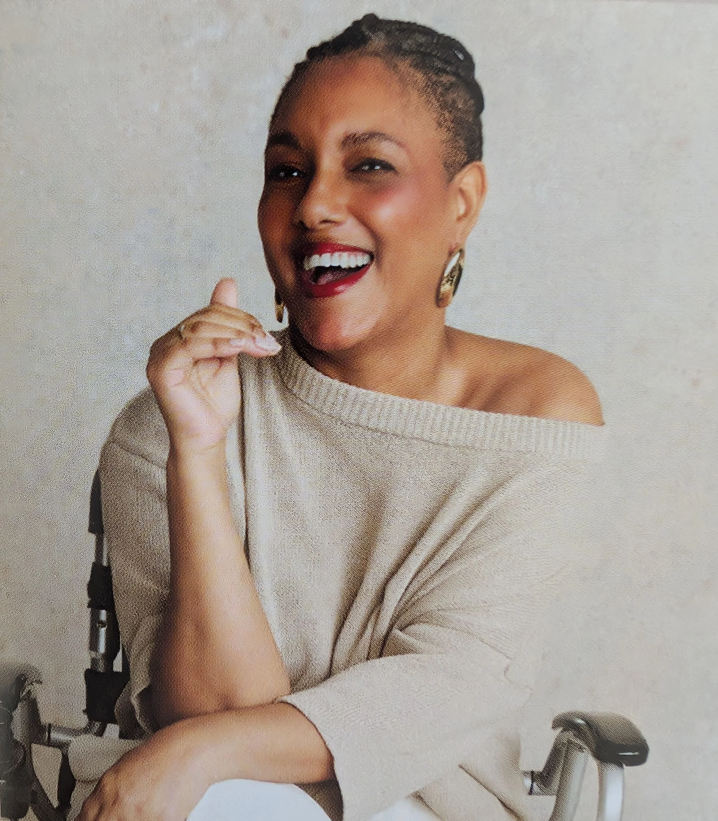 Andie is sitting in her wheelchair laughing. She wears a gold jumper off the shoulder and white trousers. Her dark braids are tied back behind her head.