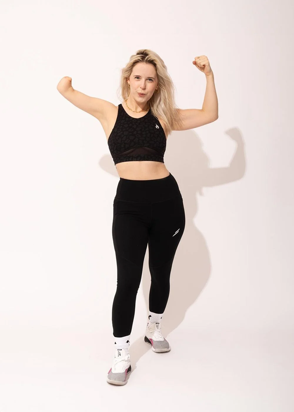 A white woman with long blond hair parted poses and flexing her muscles wearing black sports wear. She has upper limb difference in her right arm and is stood straight on looking into the camera.