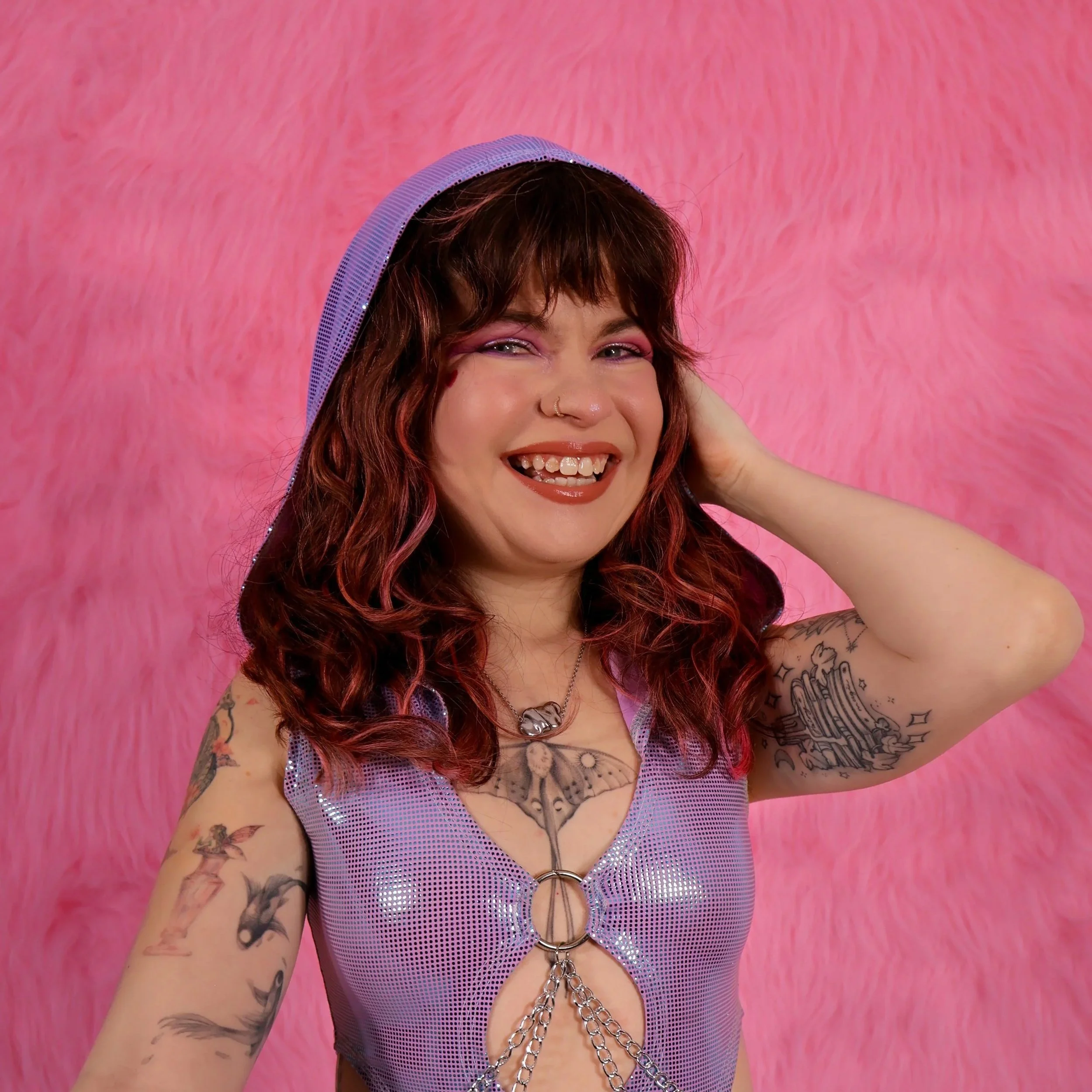 Millie is a white woman with long wavy red/brown hair. She wears a lilac body suit with a hood and pink makeup. She has tattoos on her chest and arms.