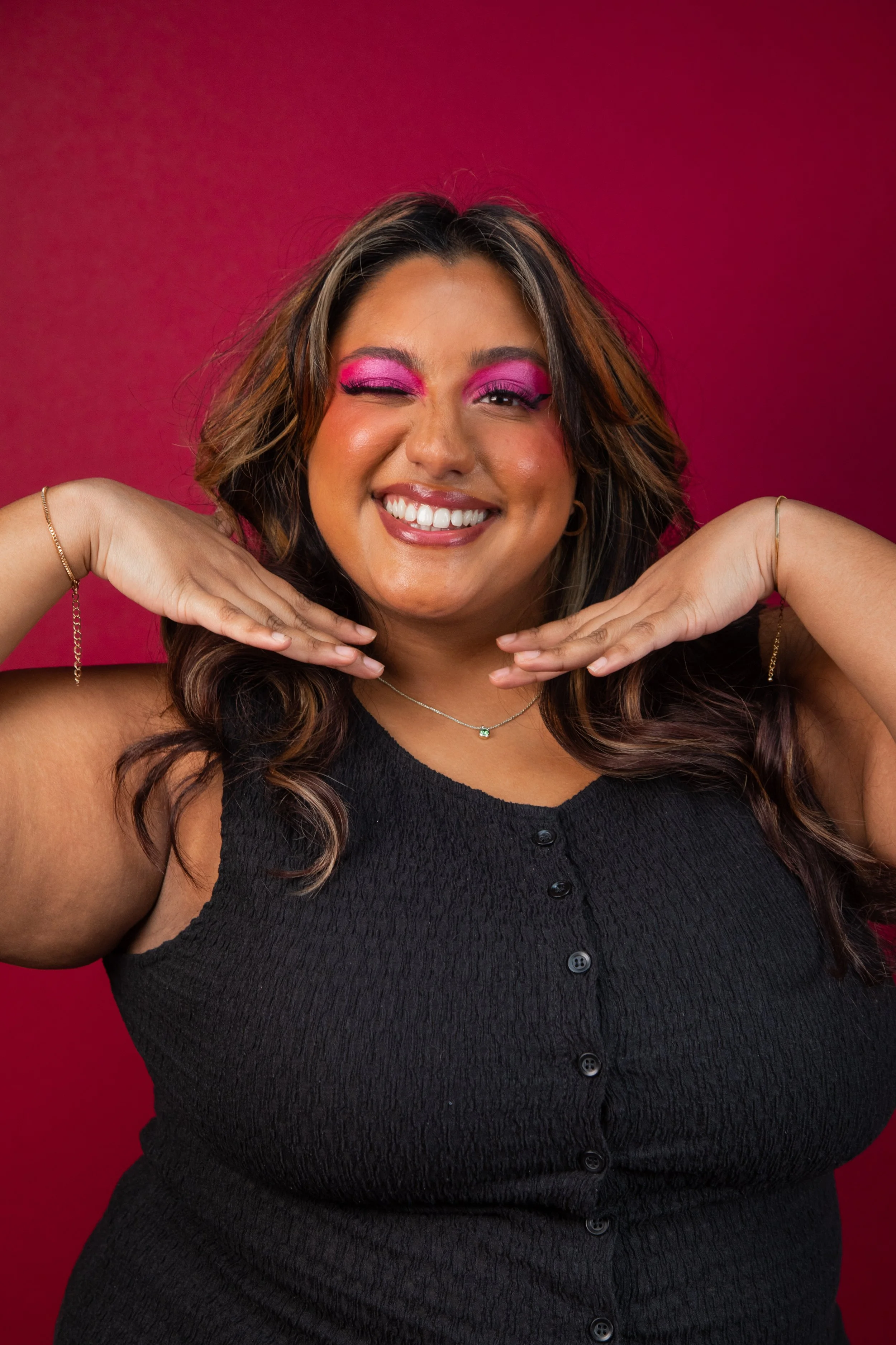 Shareena is a brown woman with long dark curly hair. Her eyeshadow is bright pink as she winks at the camera with her hands placed under her chin. She wears a black vest, stood in front of a red background.