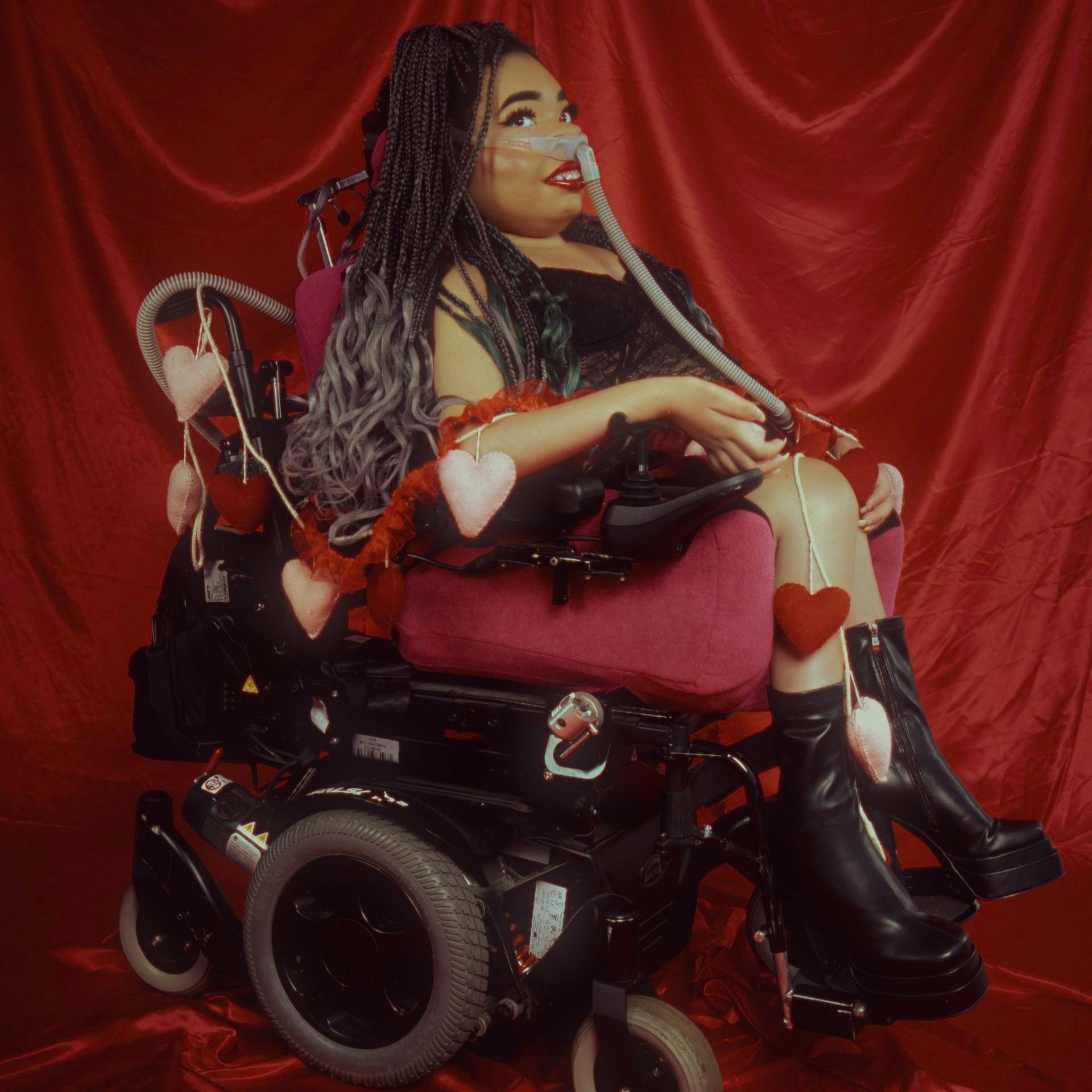 Shelby against a red velvet backdrop wearing a black dress and black boots. Her braids are dark brown and there are hearts hanging from her wheelchair.