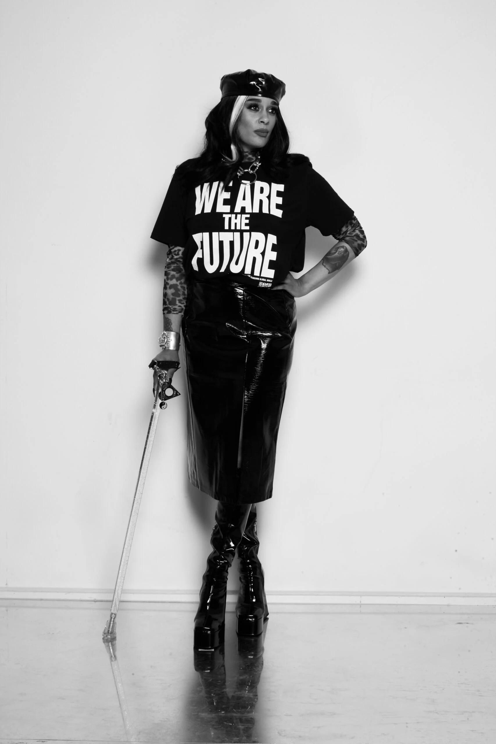 Black and White photograph of Roxy, a mixed race Disabled and pansexual femme wearing a pvc black skirt and tee with the slogan ‘we are the future’ on it. Standing with a walking stick, they are wearing a wearing a beret over their long dark hair. 