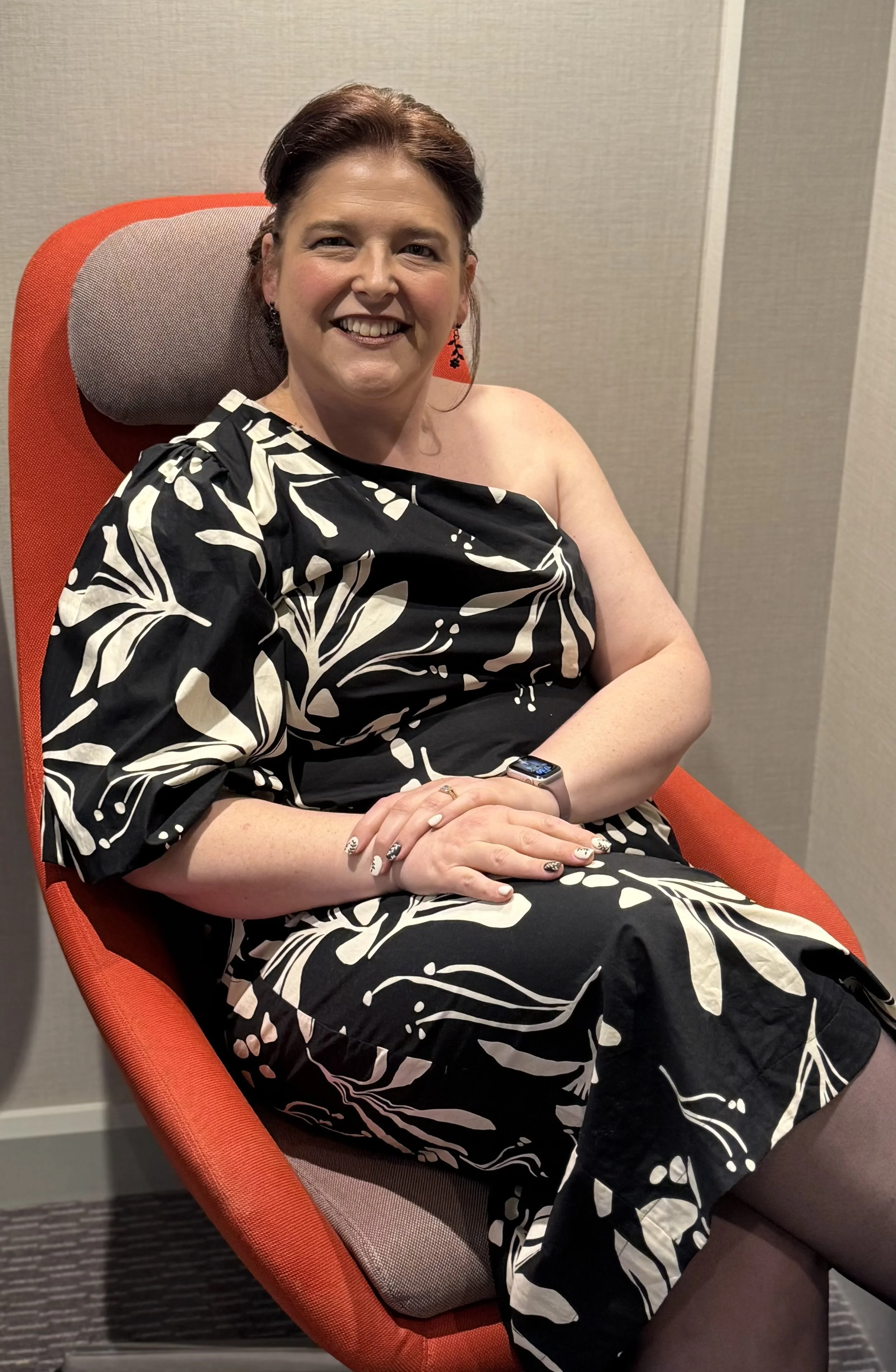 Deanne is sitting in an orange chair with a grey headrest. Her hair is brown and pulled back and she wears a black and white off the shoulder dress. Her legs are crossed and her hands sit on her lap as she smiles.