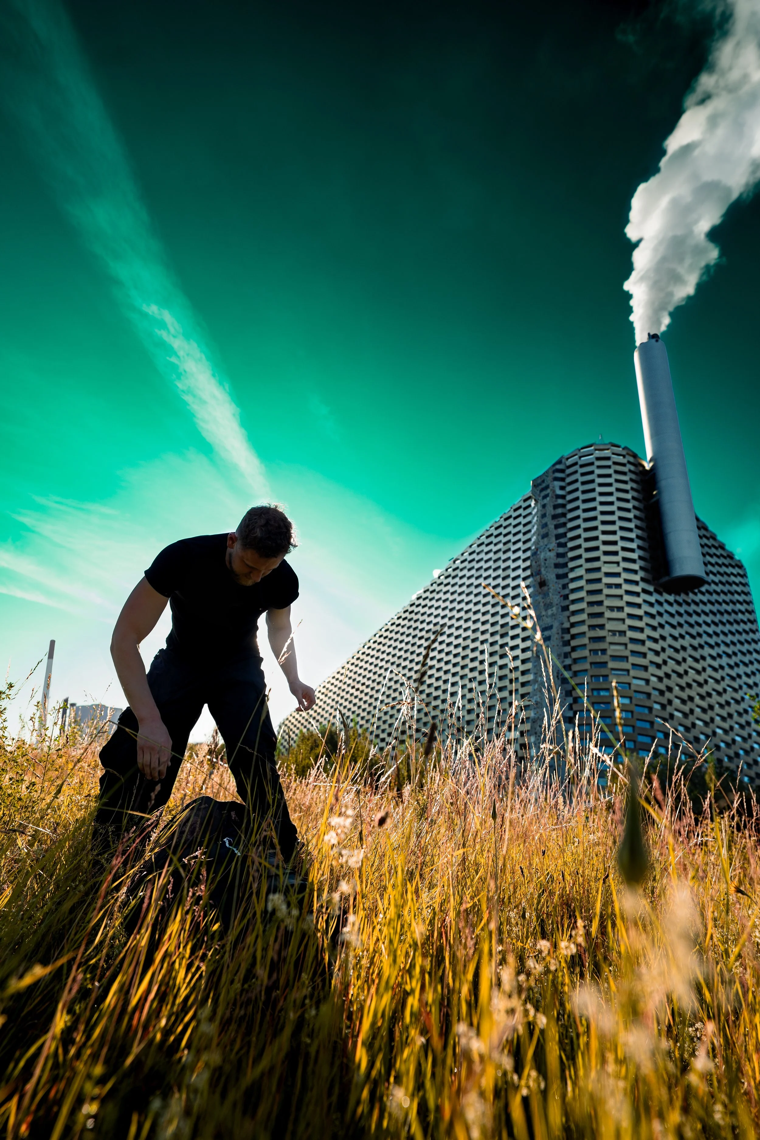George is stood looking down at long grass. He wears and black t.shirt and black trousers in front of Copenhill. The sky is a saturated green colour.