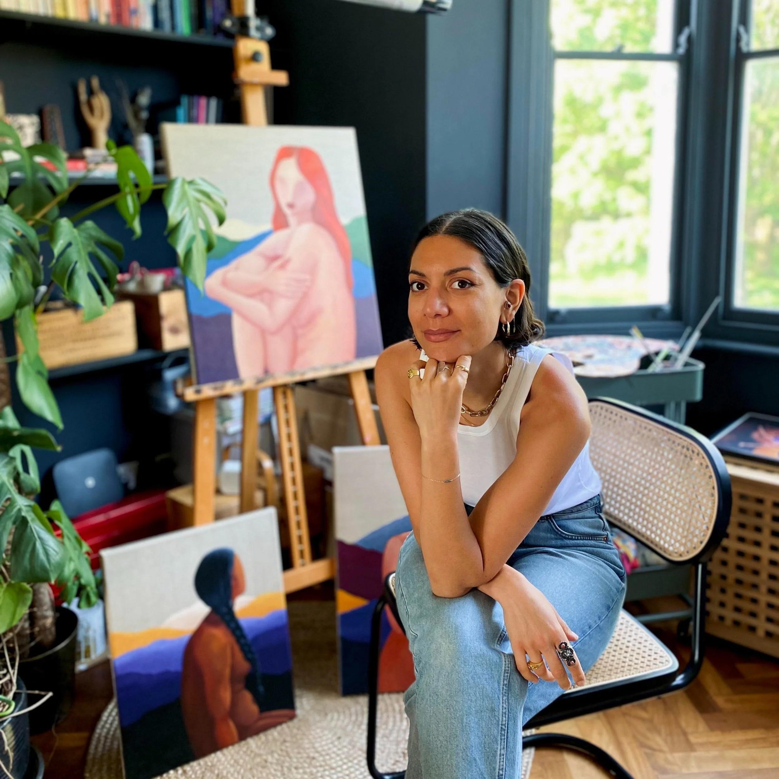 Myriam is a mixed race woman with brown hair sitting in her studio. There is a painting of a woman on an easel in the background.