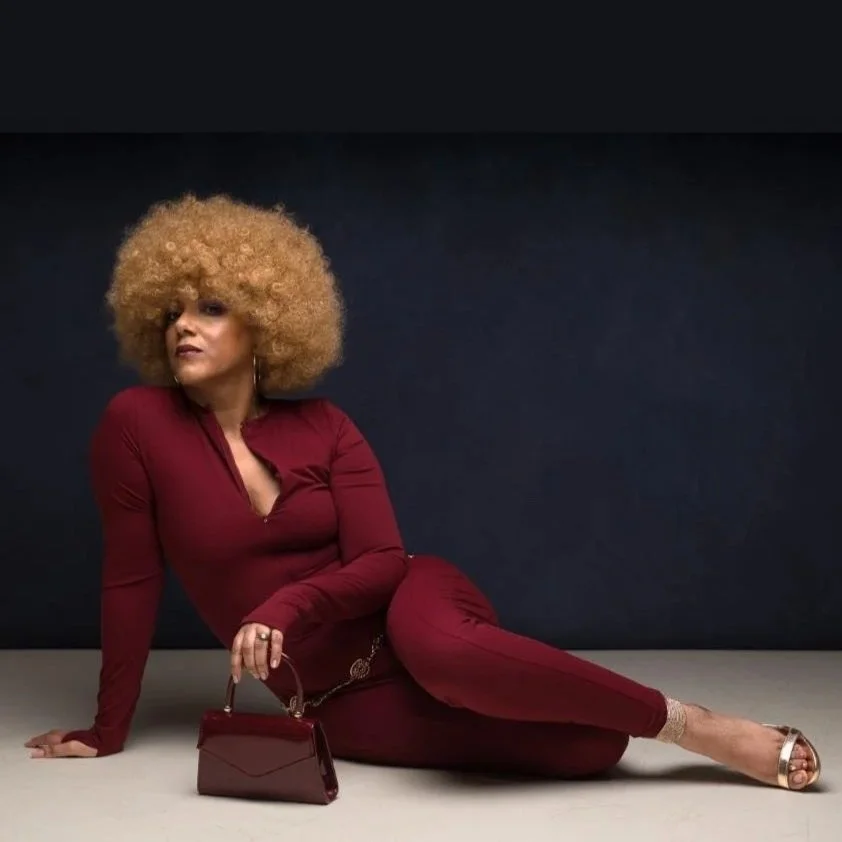 Andie is a black woman with a short blonde tight afro. She wears a burgundy jumpsuit and is sitting on the floor, posing gracefully with her legs to one side.