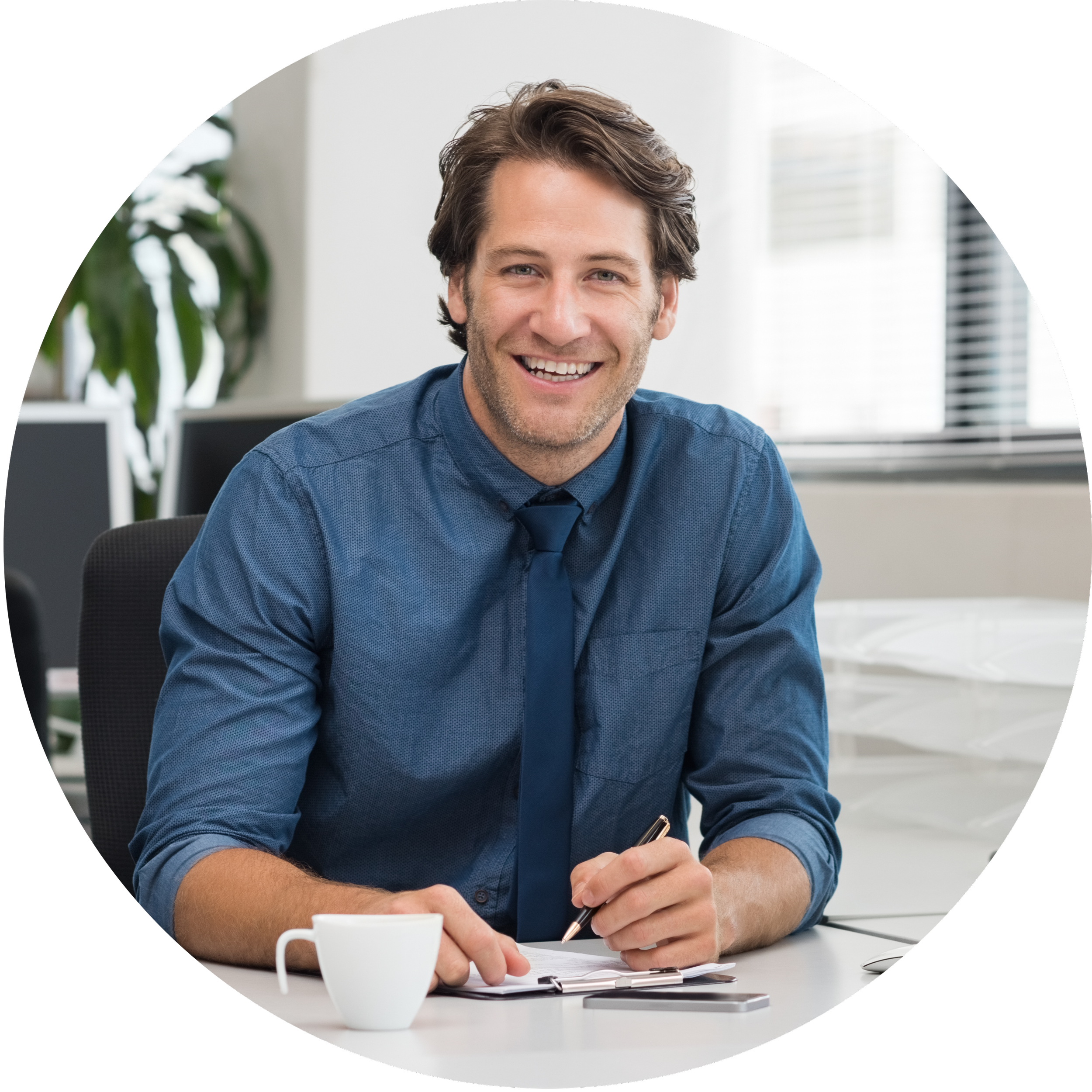 Man in blue shirt and tie smiling while sitting at a desk in an office, with a notepad, pen, coffee cup, and smartphone.