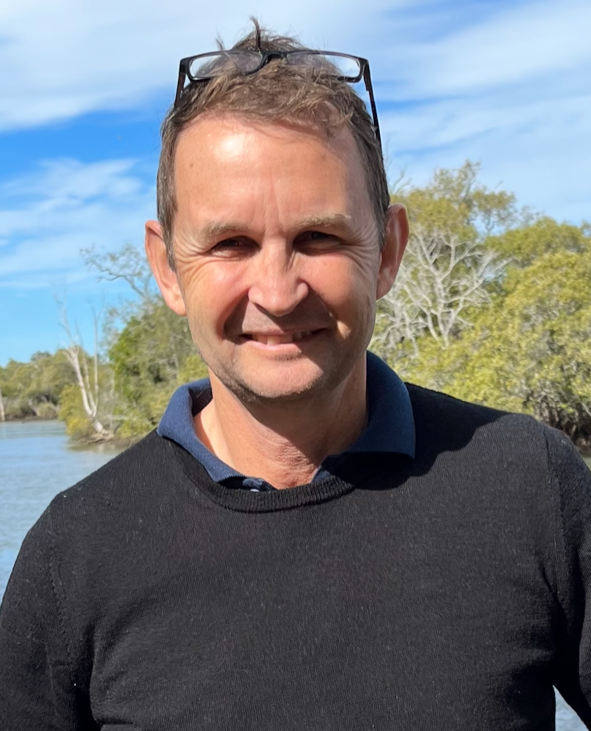 A man outdoors near a river with greenery and trees in the background, wearing a dark sweater over a collared shirt, with glasses on his head, smiling at the camera.