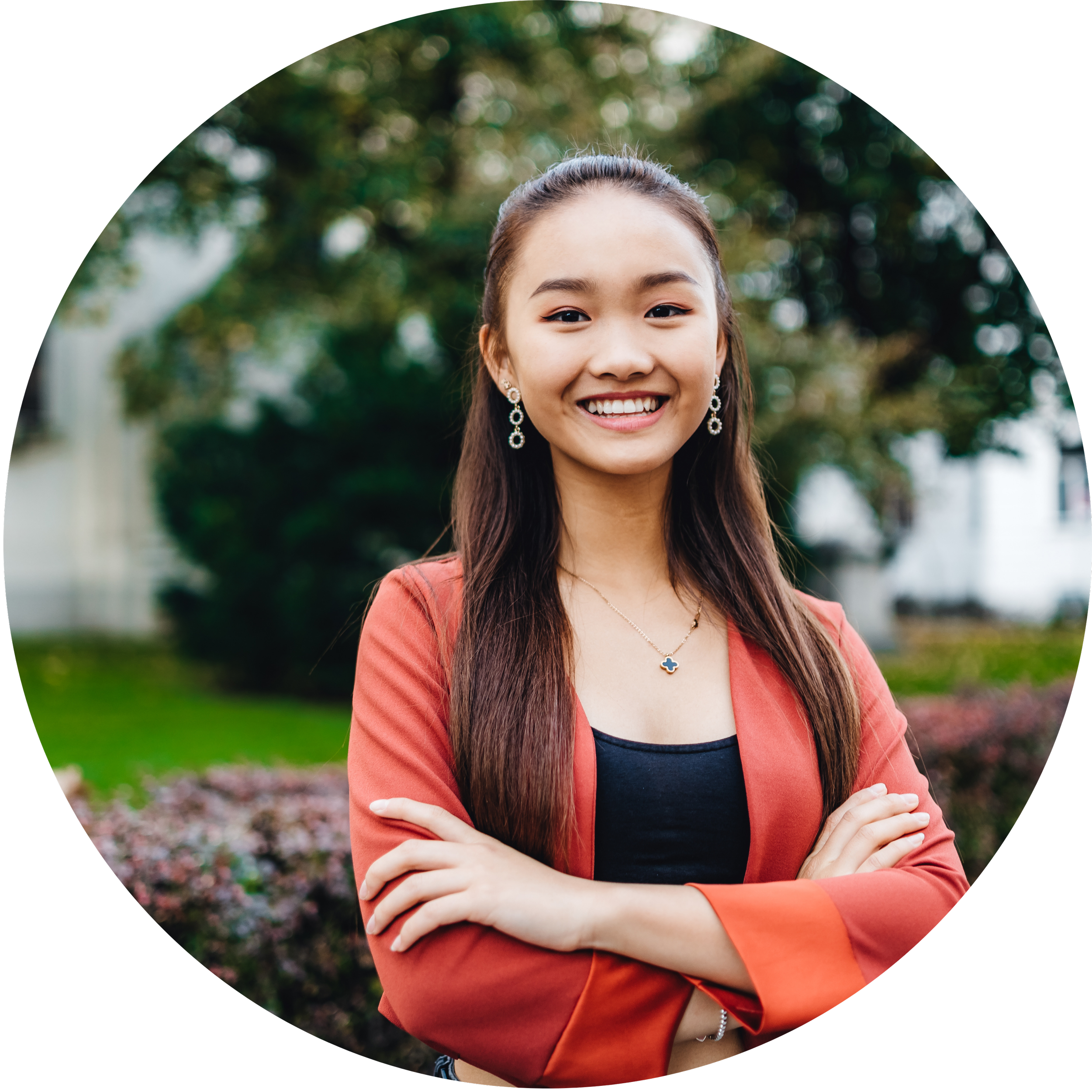 A young woman smiling outdoors, with arms crossed. She has long dark hair, wears earrings and a necklace, and is dressed in a black top with an orange blazer. Background features trees and a white building.