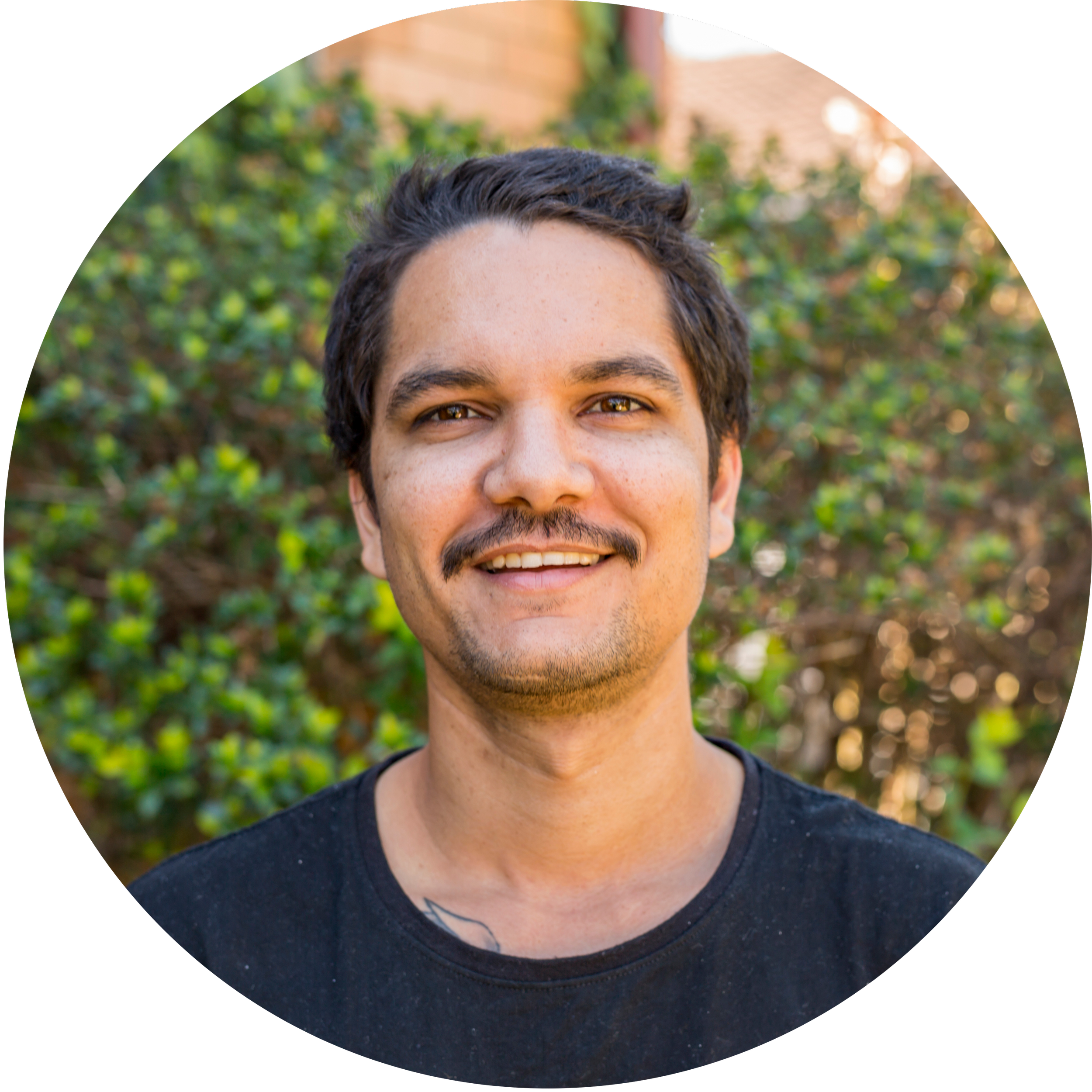 A young man with dark hair, a mustache, and a goatee smiling outdoors with a background of green leafy bushes and a brick building.