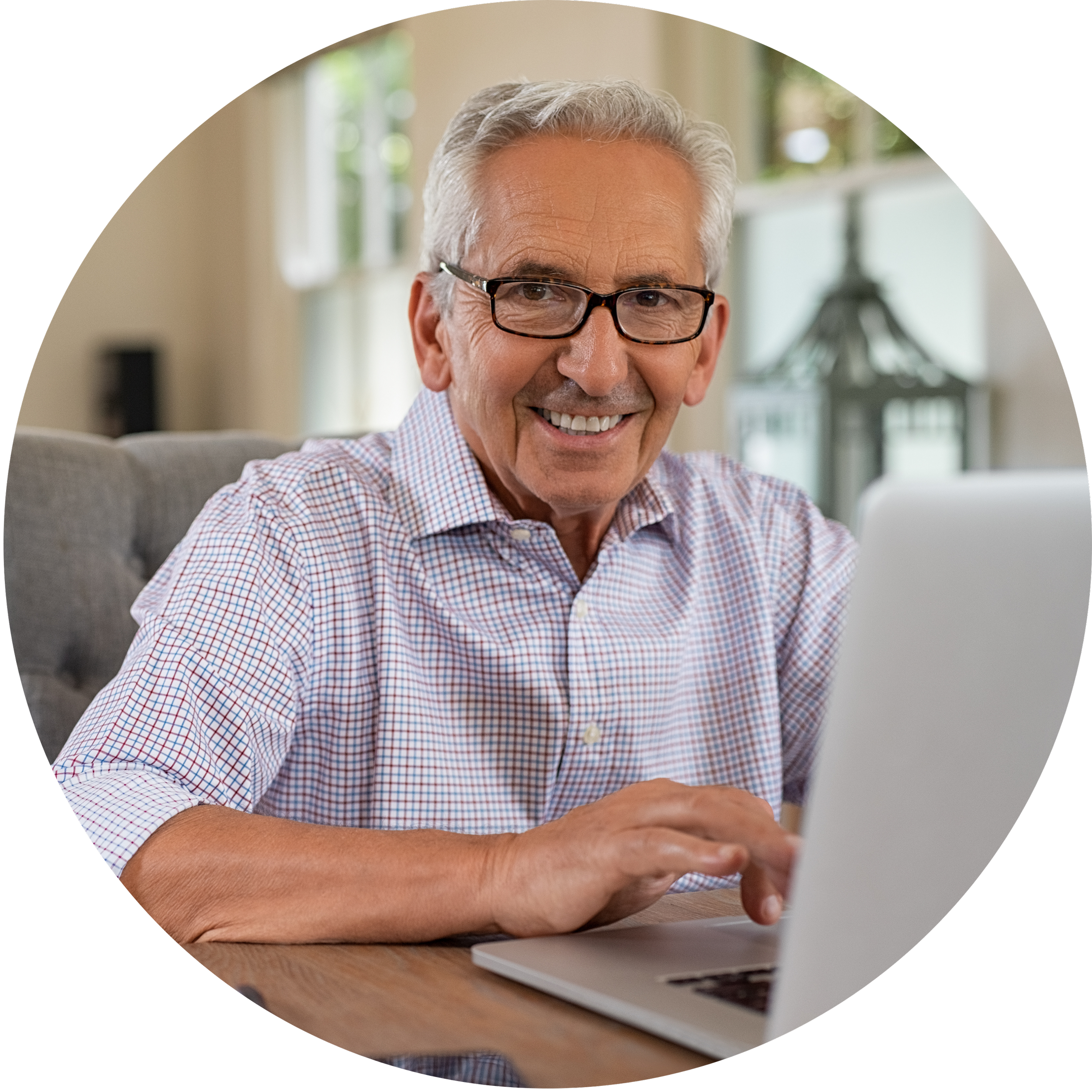 An older man with glasses, gray hair, wearing a checkered shirt, smiling while using a laptop at home.