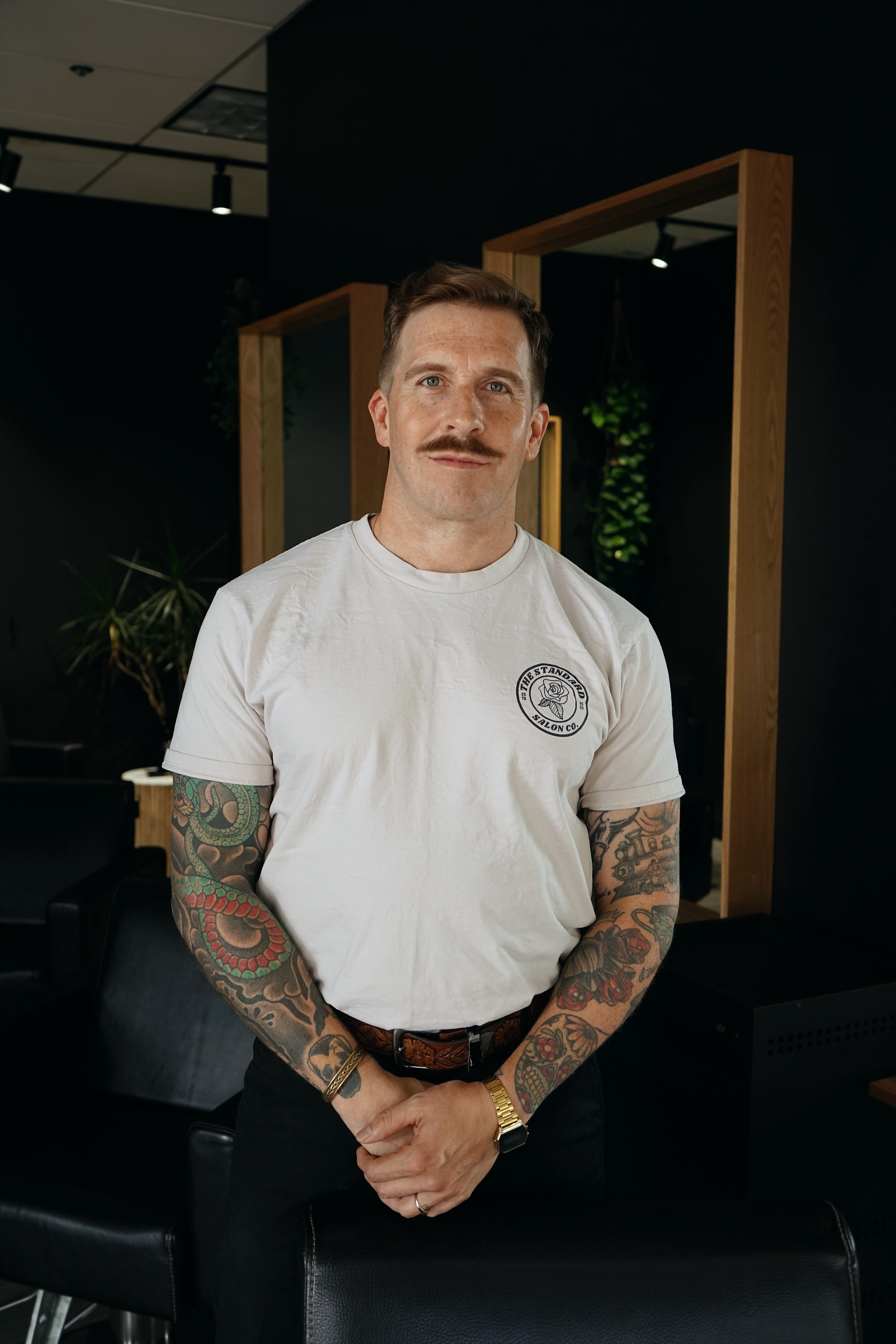 A man with long hair and tattoos wearing a white T-shirt with 'Campfire Smores' logo, holding black clothing, standing in a room with a mirror and plants.