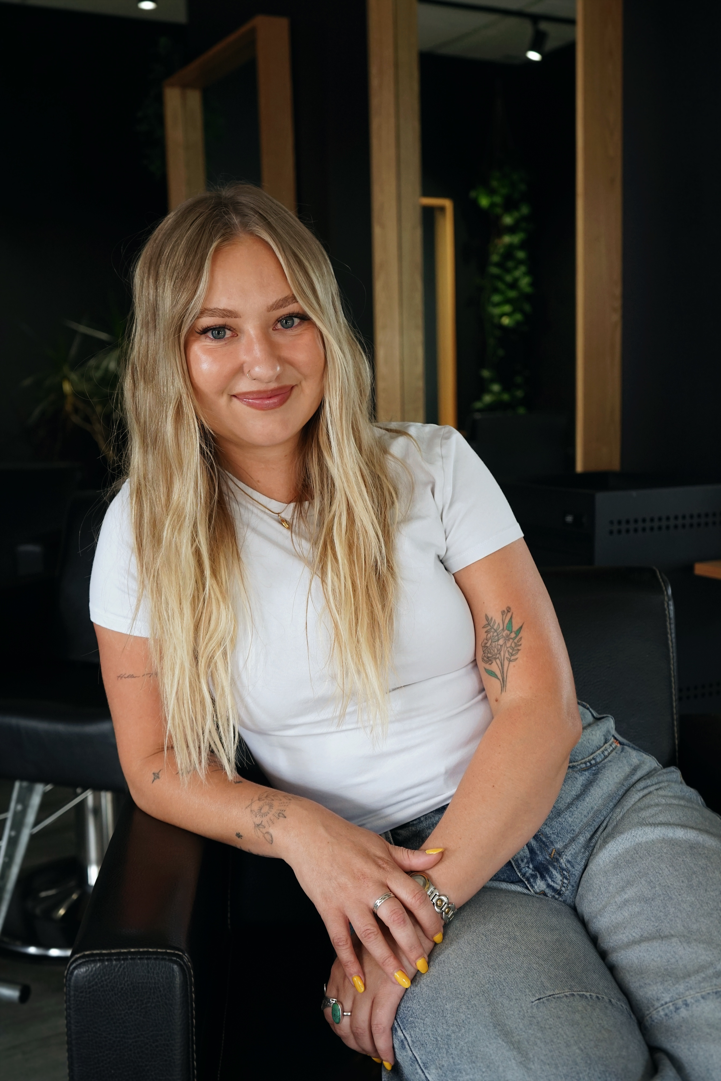 A woman with light brown hair sitting on a beige cushioned chair, wearing a black tank top and light blue jeans, with a black watch and rings, in a room with beige walls and dried plants.