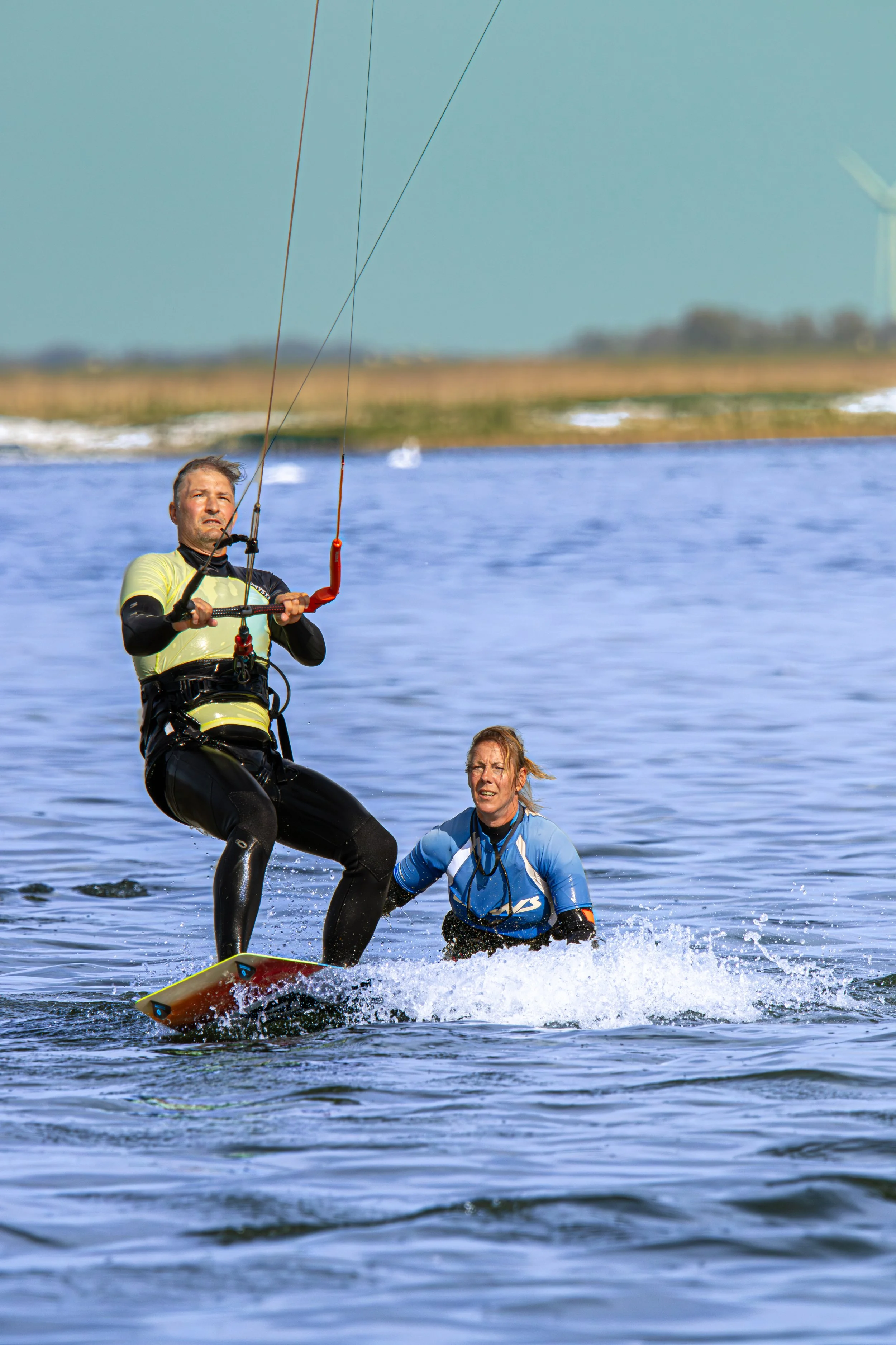 Man die standup paddleboard bestuurt terwijl een vrouw in het water naast hem zit, beiden op open water met windmolens op de achtergrond.